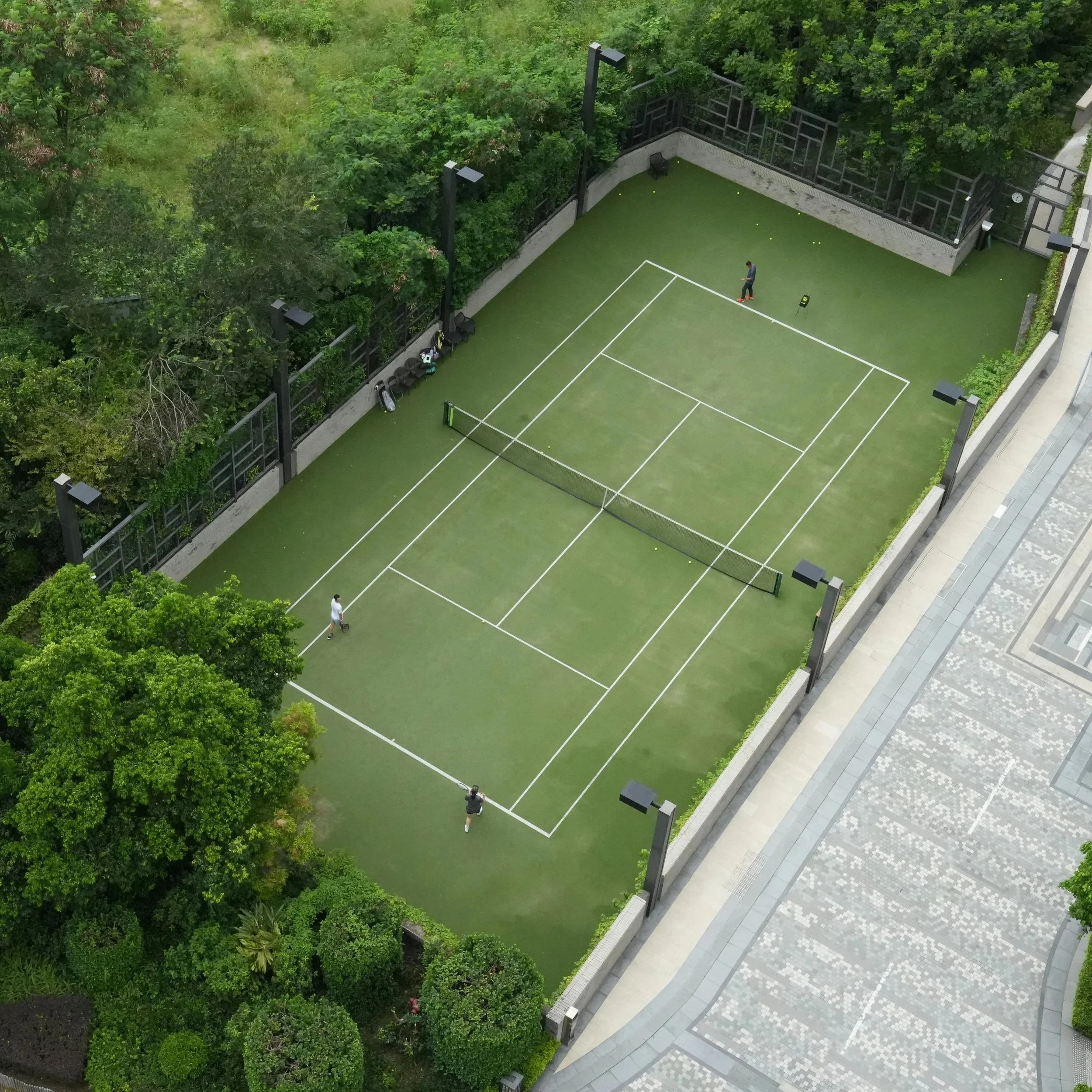 An aerial view of a small tennis court surrounded by lush green trees and bushes, with a person standing on the court and another nearby.