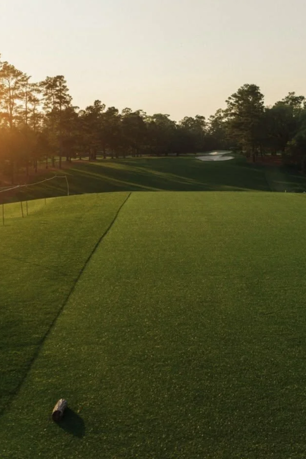A golf course at sunset with a tee box and a line marking the edge of the fairway, surrounded by trees and a sand trap in the distance.