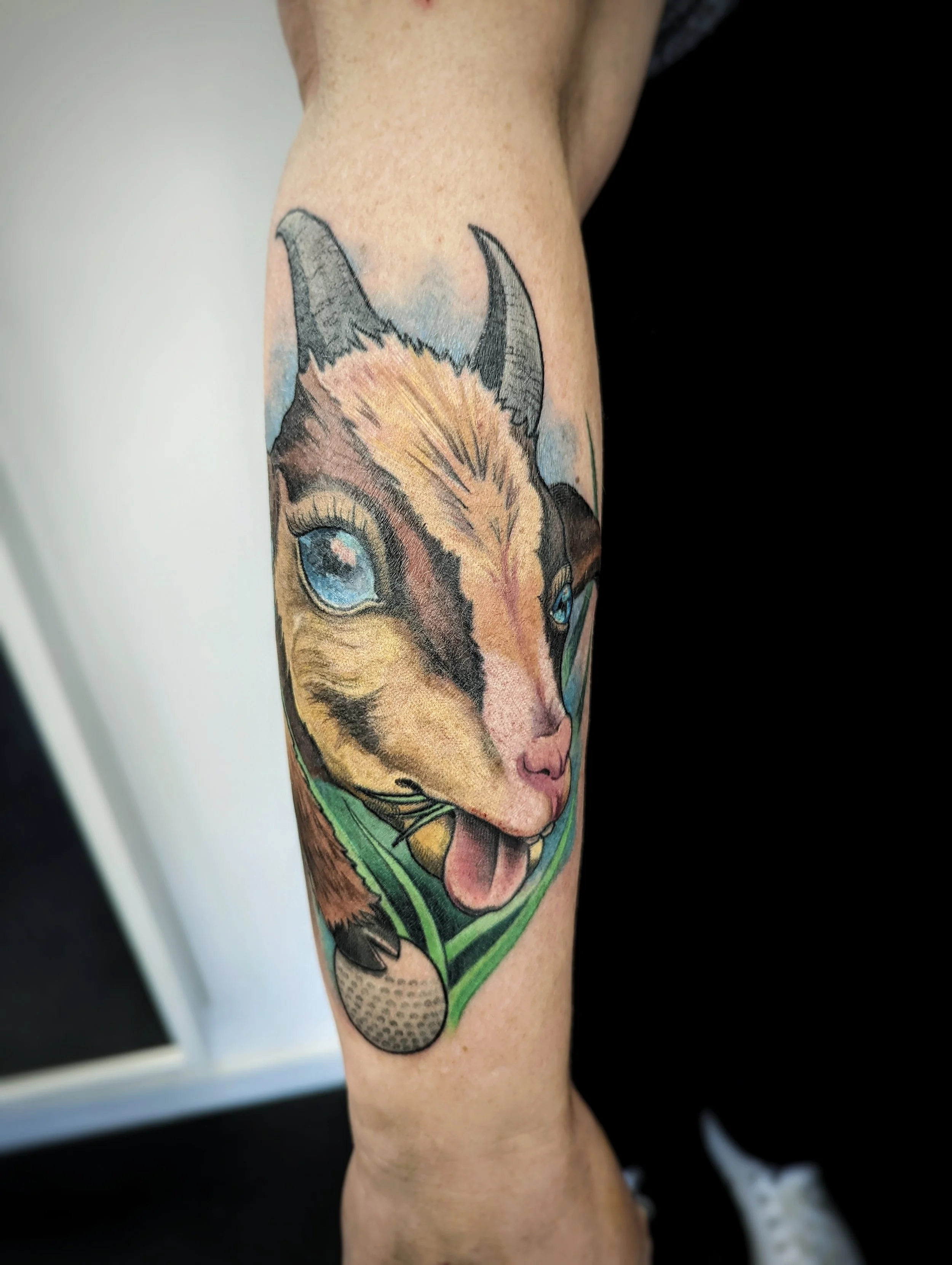 Tattoo of a dog's face on person's arm, with detailed fur, blue eyes, and tongue sticking out, surrounded by green leaves.