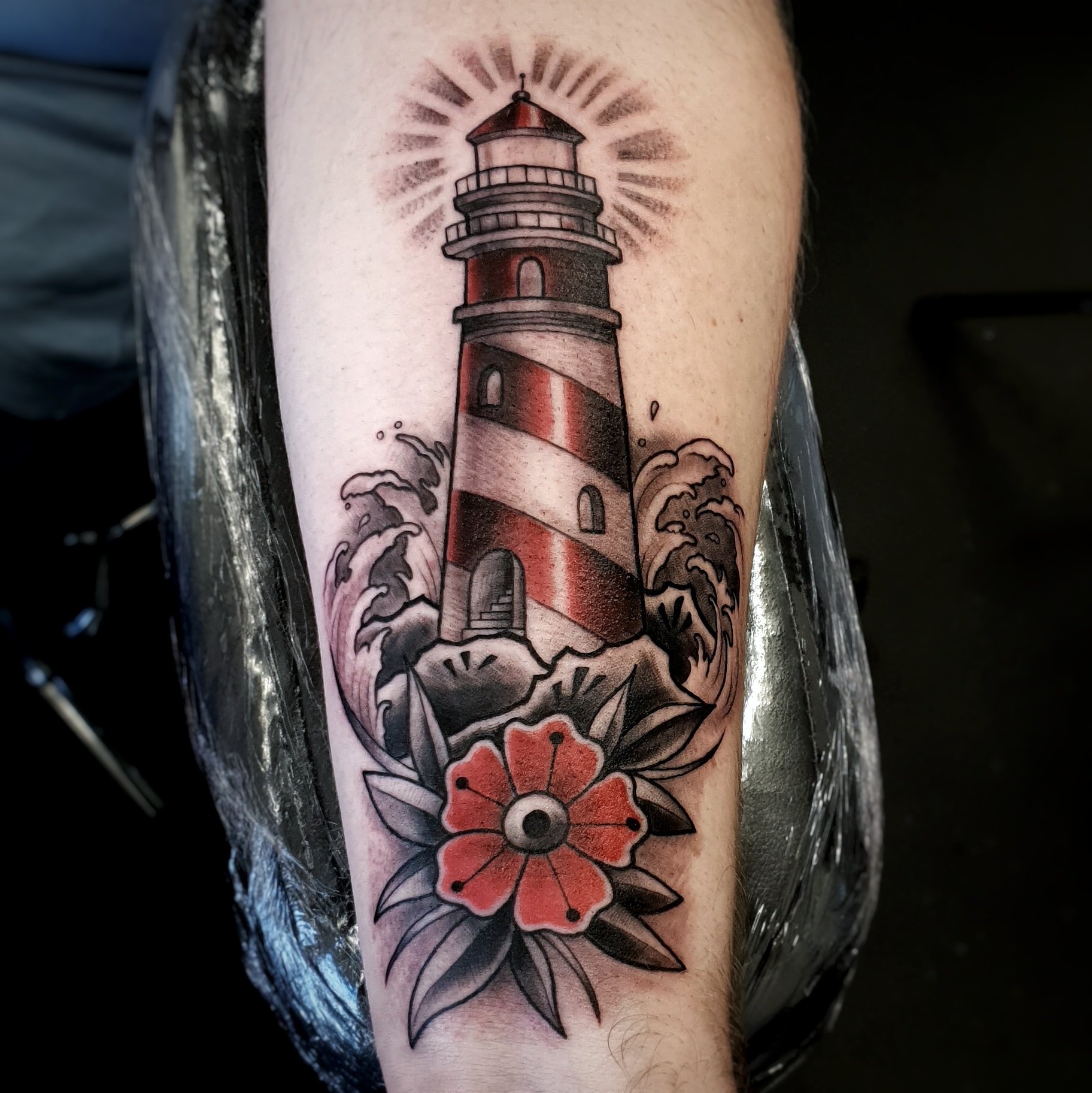 Tattoo of a lighthouse emitting rays of light, with ocean waves at the base and surrounding floral elements.
