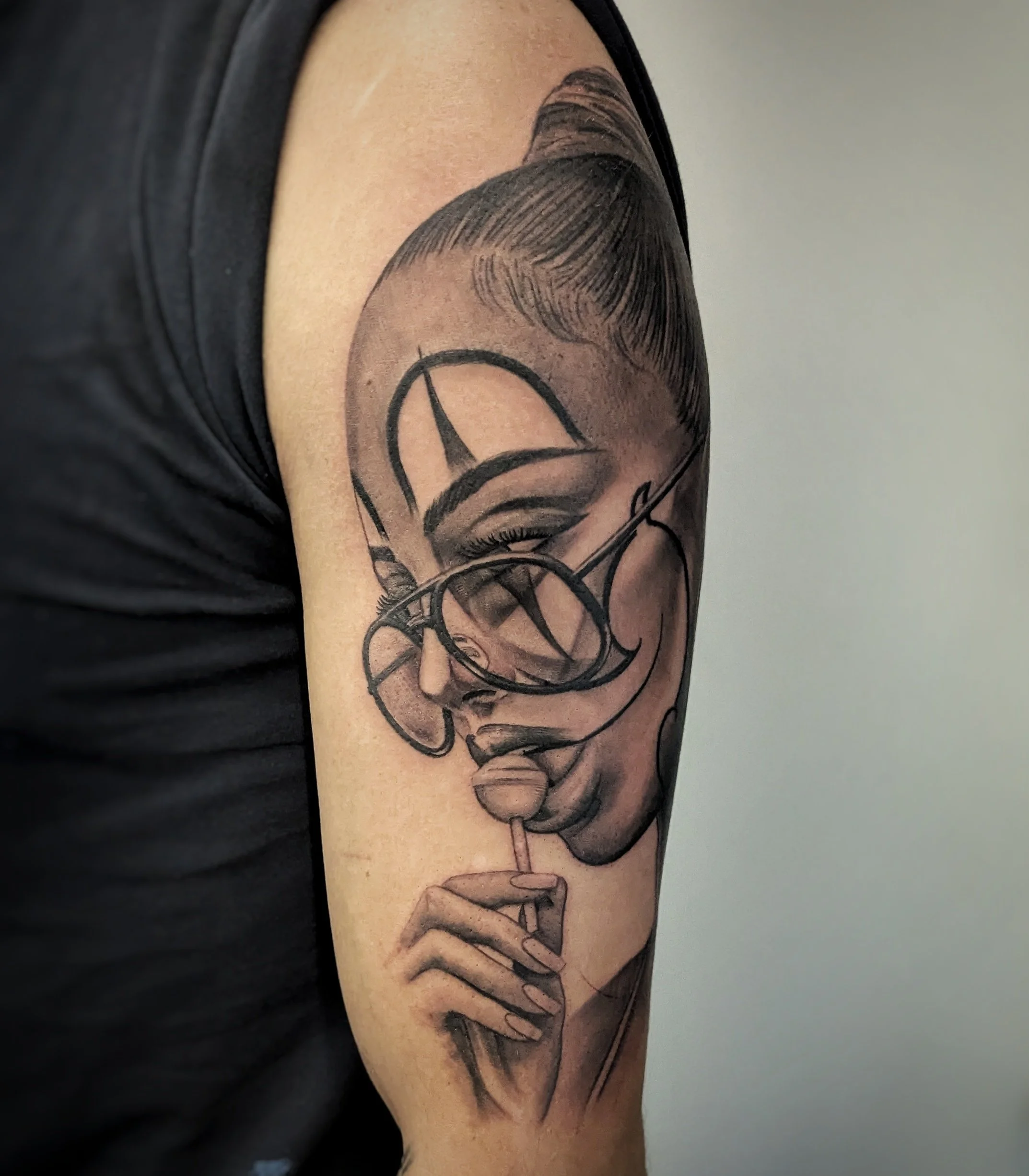 A person's upper arm with a black and grey tattoo of a woman's face wearing glasses, with wavy hair and closed eyes.