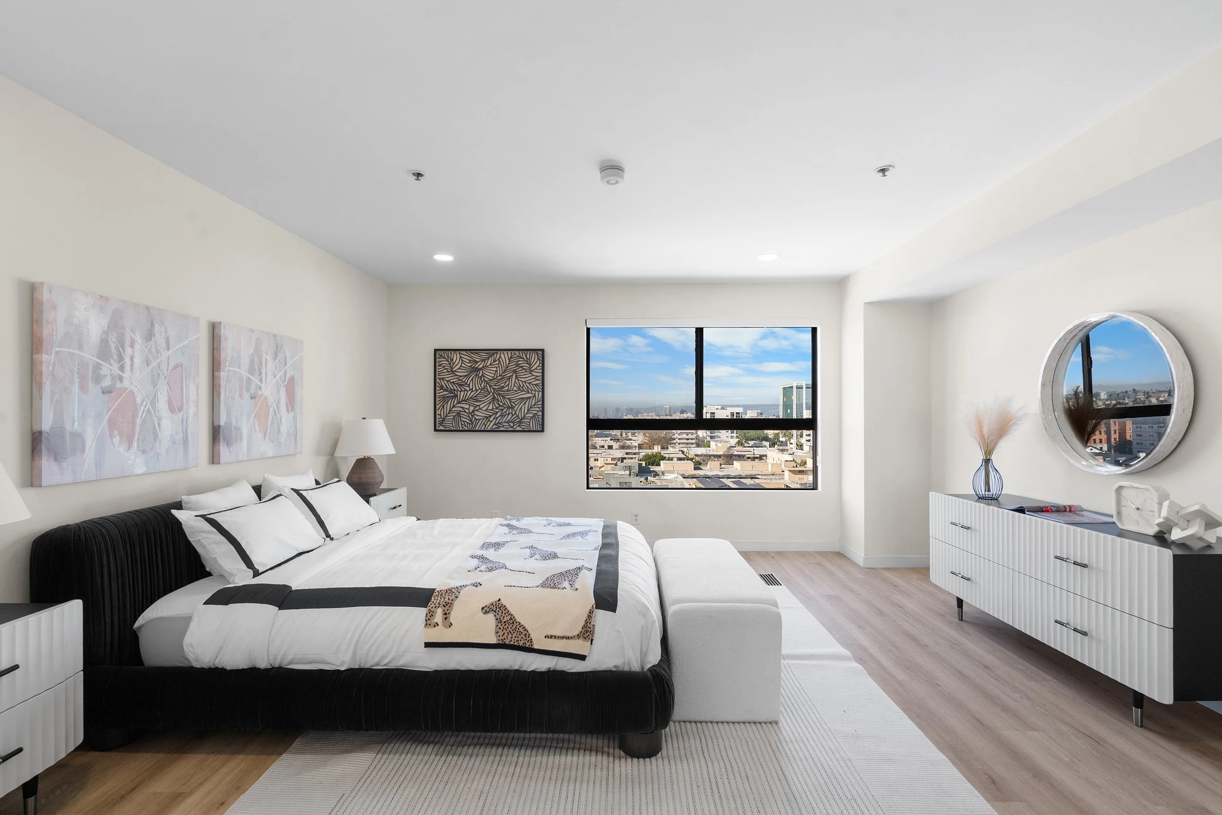 Modern bedroom with white walls, large window showing city view, black bed with white bedding and animal print blanket, abstract artwork, white dresser with decorative objects, and round mirror.
