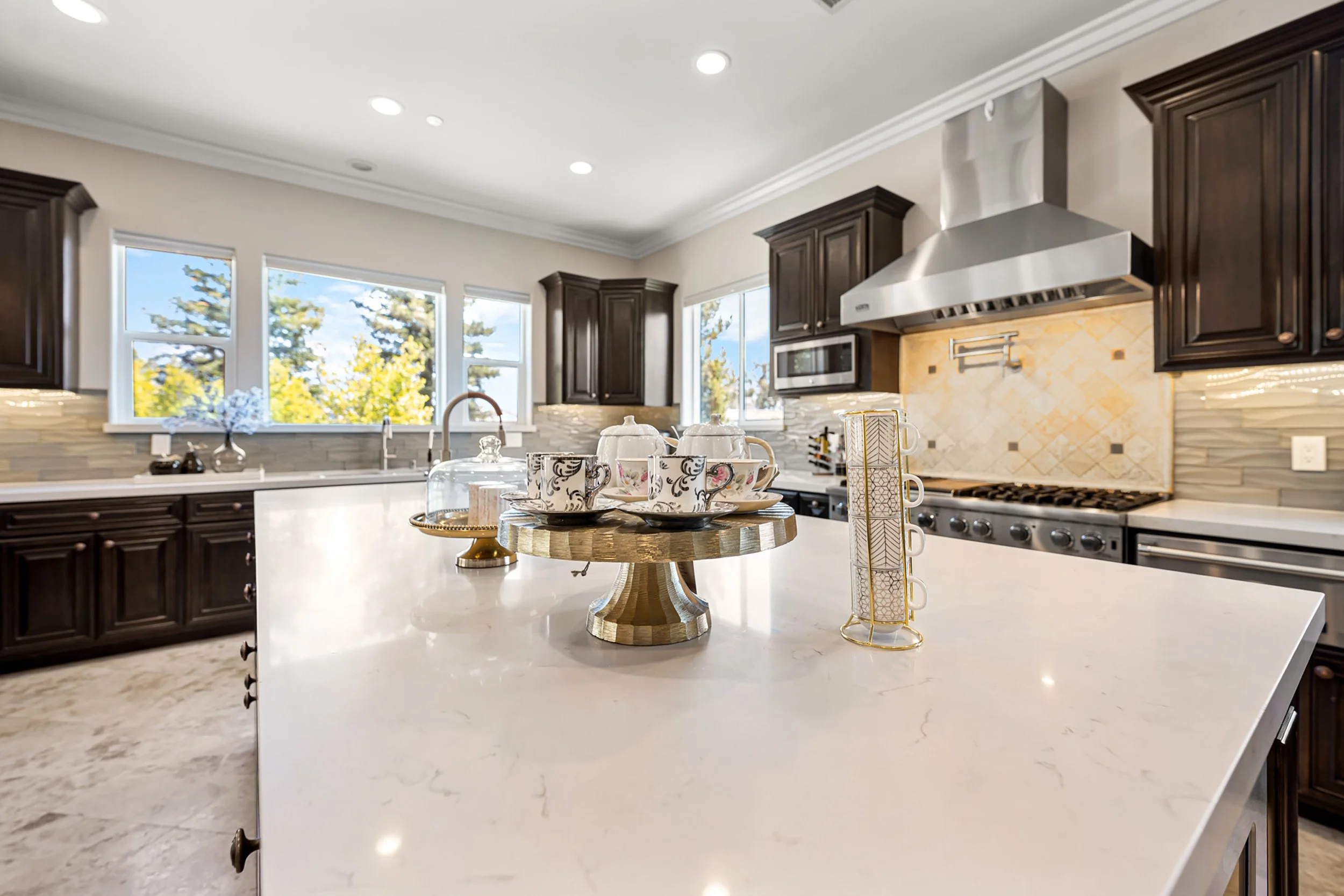 Modern kitchen with dark wooden cabinets, stainless steel range hood, countertop with decorative cups on a wooden cake stand, and large windows showing trees outside.