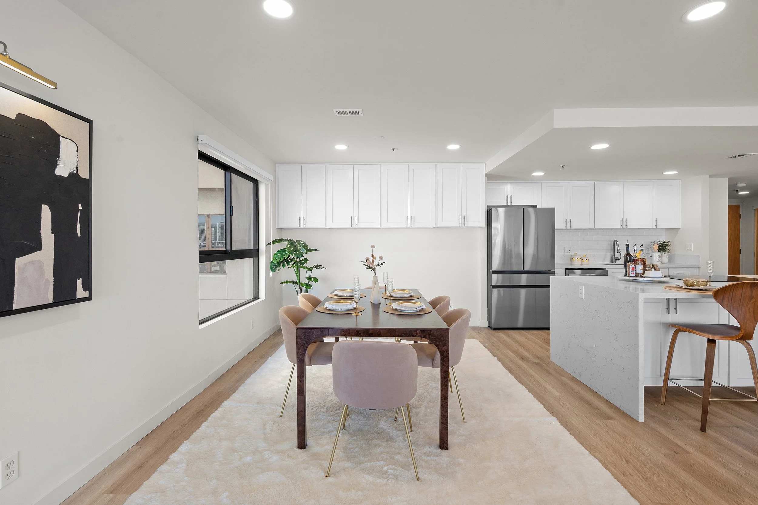 Open-concept kitchen and dining area with white cabinets, stainless steel refrigerator, dining table set for four, beige chairs, decorative plants, and a large black and white abstract art piece on the wall. Recessed lighting and a window with a city