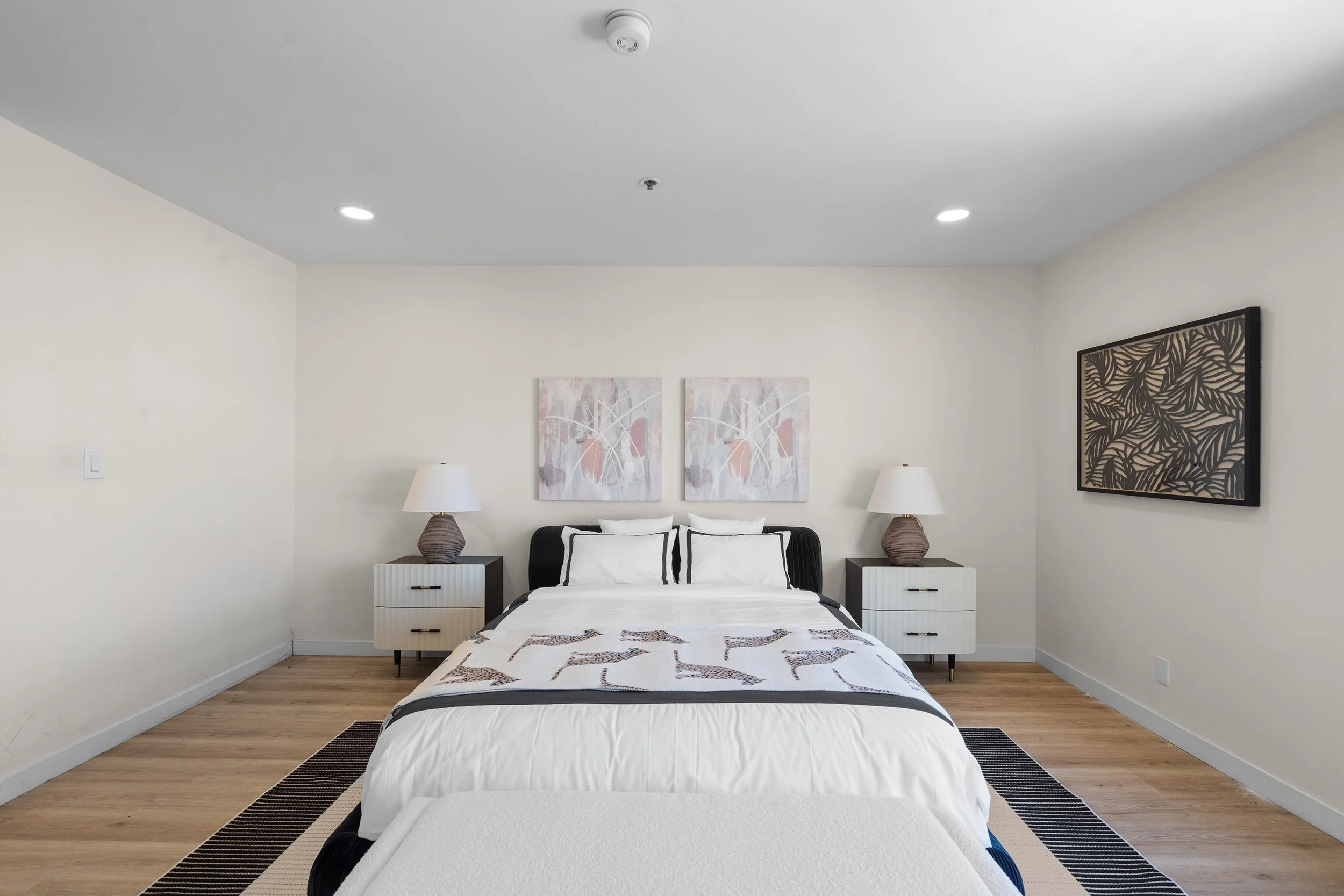 A bedroom with a bed, two nightstands with lamps, decorative artwork on the walls, and hardwood floors.
