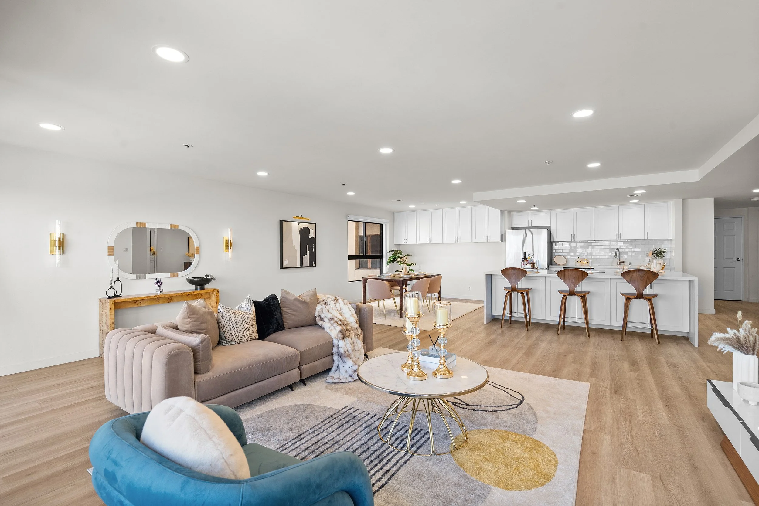 Modern open-concept living room and kitchen with white walls, light wood flooring, beige couch with pillows, a blue velvet chair, a coffee table with gold accents, and a white kitchen with bar stools and a subway tile backsplash.