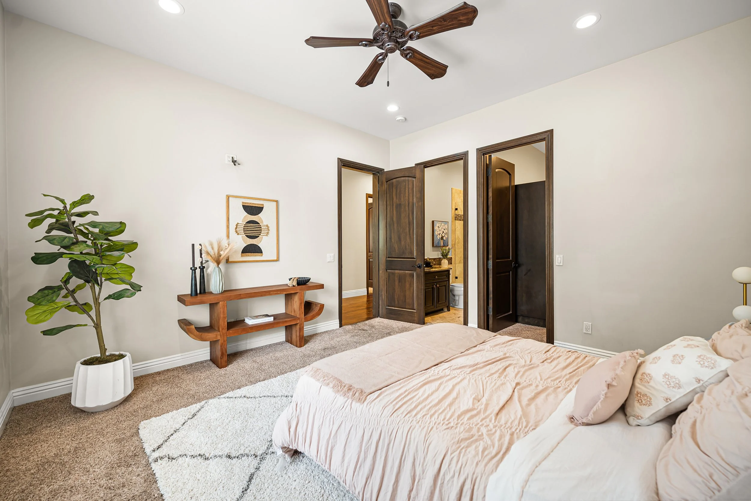 A bedroom with a bed covered in a light pink quilt and pillows, a large potted plant in a white pot, a wooden console table with decorative vases and a book, framed abstract art on the wall, open dark wooden doors leading to a bathroom with a vanity 