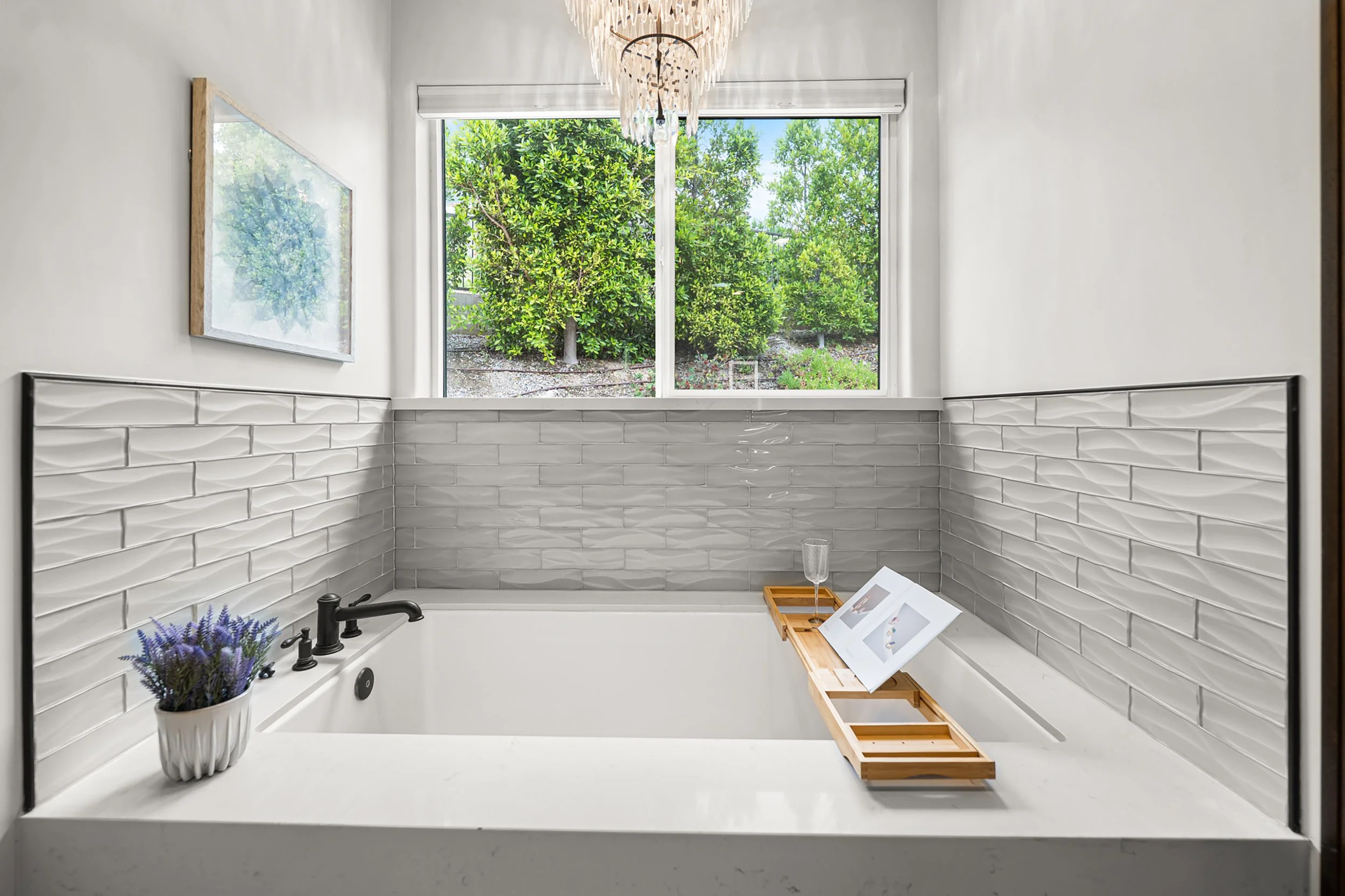 Bathroom with large window showing greenery outside, white tiled walls, a small tray with a wine glass, and a purple flower plant.