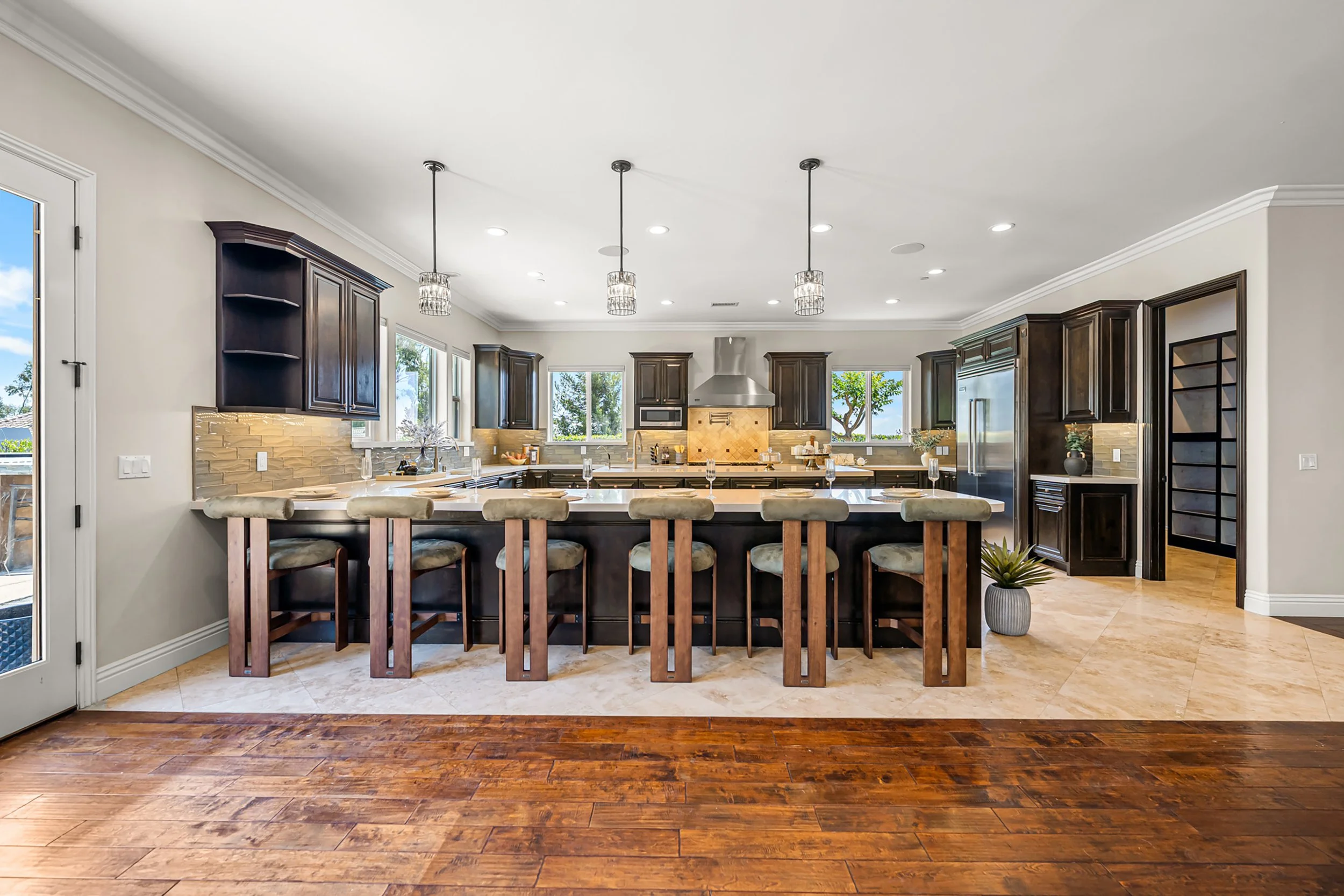 Bright modern kitchen with dark wood cabinets, beige tile backsplash, granite countertops, stainless steel appliances, and large windows with a view of trees.