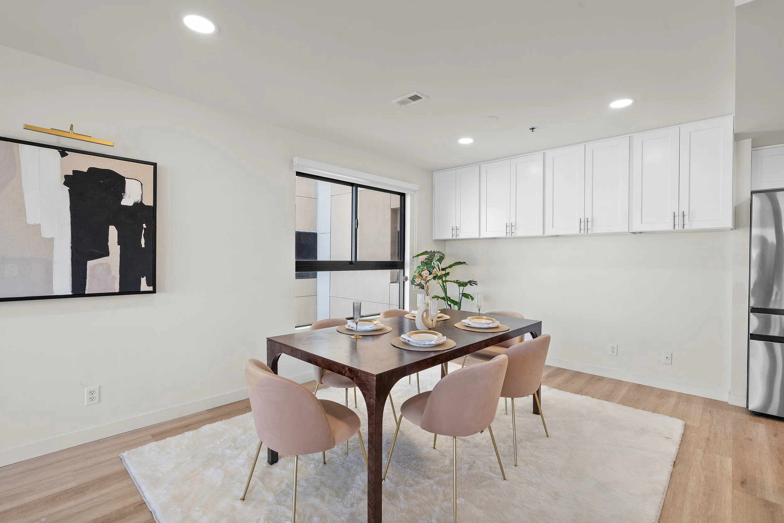 Interior of a modern dining area with a wooden dining table, pink upholstered chairs, a white rug, and a black window. There are white kitchen cabinets and a stainless steel refrigerator. A plant with pink flowers is on the table, and a large abstrac