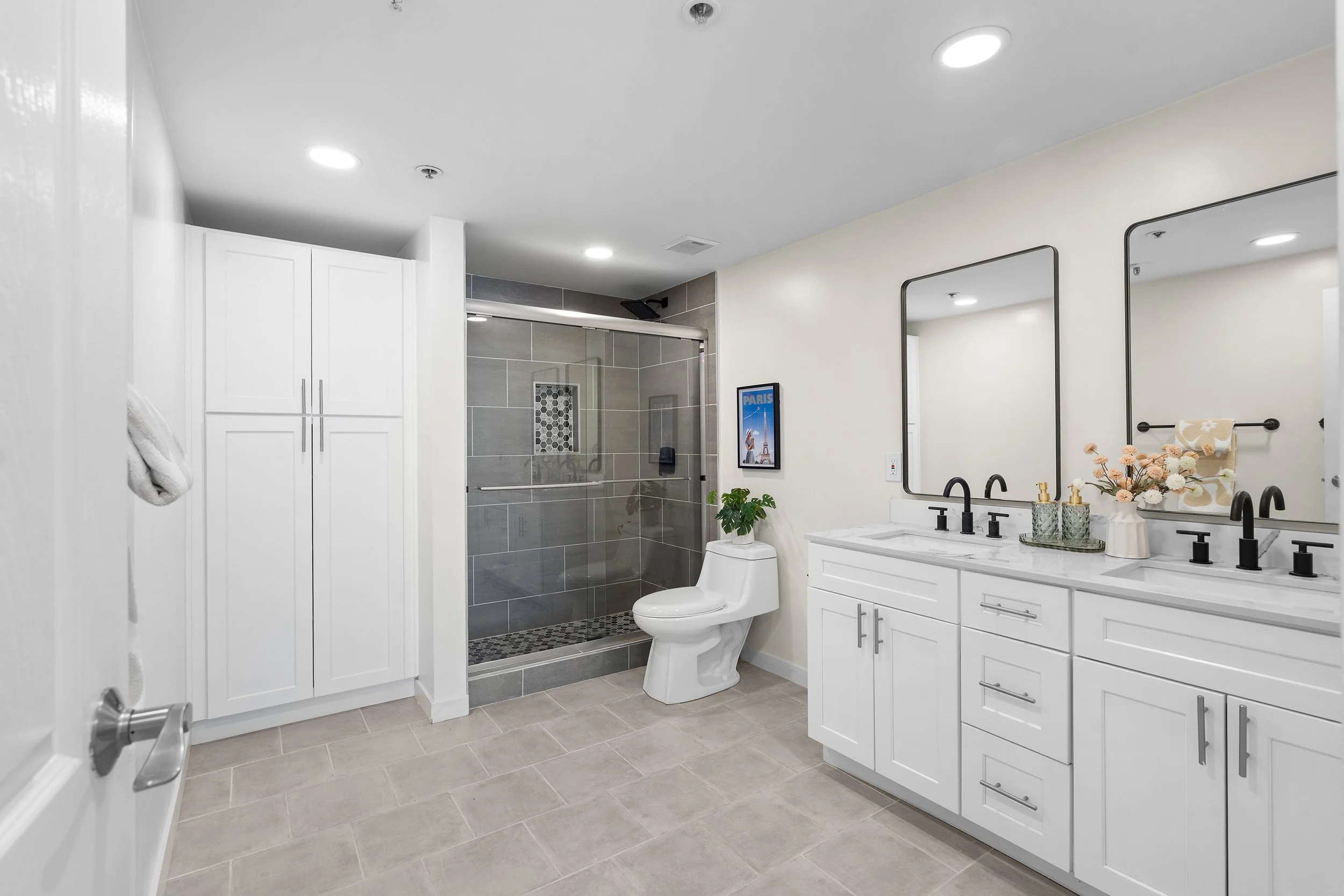 A modern bathroom with a double vanity, large mirrors, a toilet, and a walk-in shower with gray tiles and a glass door.