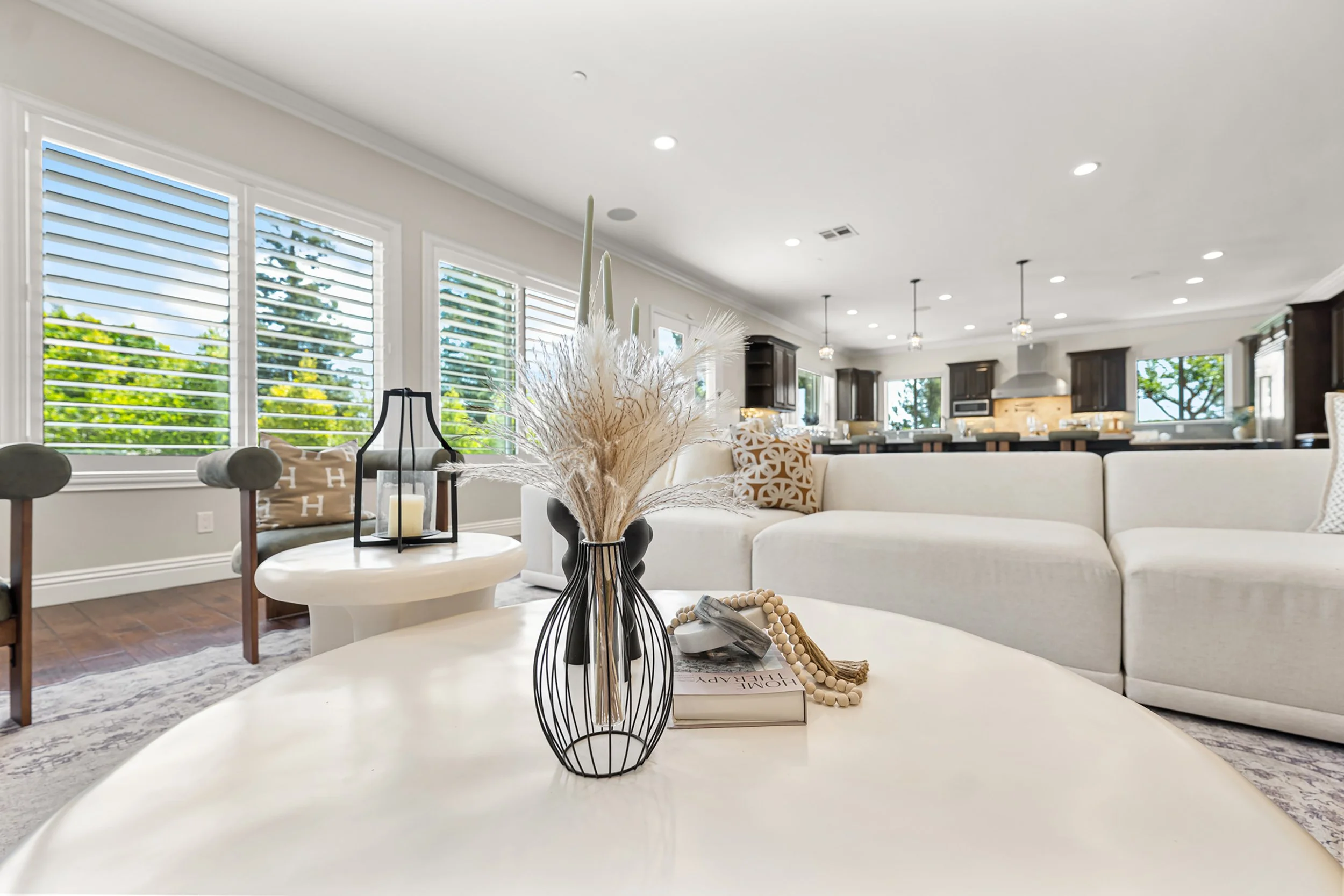 Bright living room with white walls, large windows with white shutters, and a white sectional sofa. Decor includes a black vase with dried pampas grass on a white table, and pillows on the sofa. Kitchen area with dark cabinets and a large range hood 