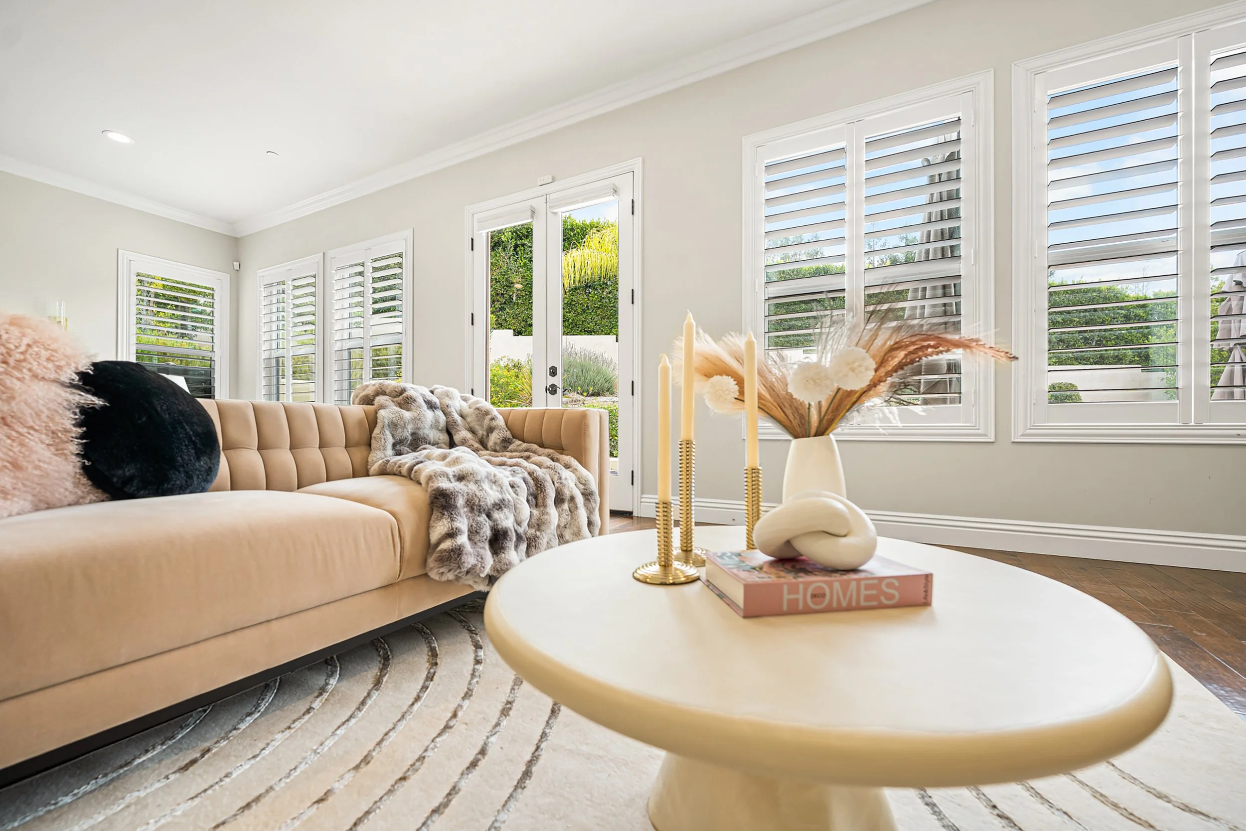Living room with beige sofa, decorative pillows, and a coffee table with candles and a flower arrangement. Large windows with white shutters and a view of a garden outside.