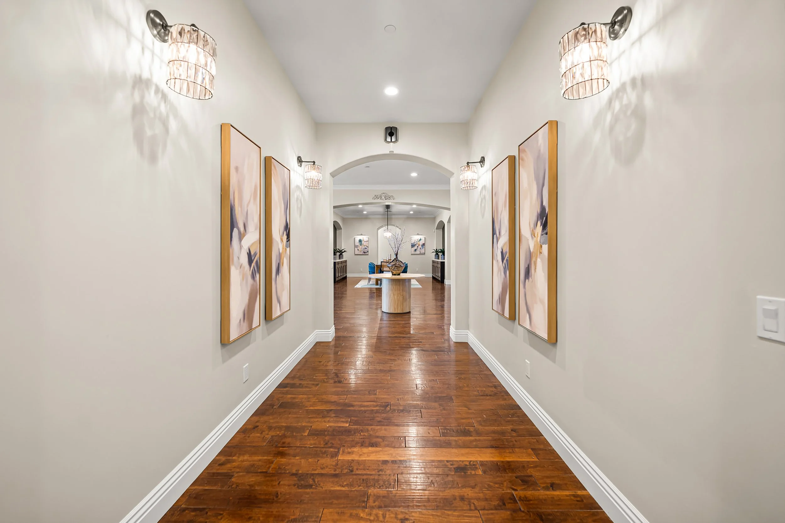 A hallway with white walls and wooden floors, decorated with four paintings framed in wood, two on each side, and two wall-mounted light fixtures that cast shadows on the walls. The hallway leads to a dining area with a round table, a flower arrangem