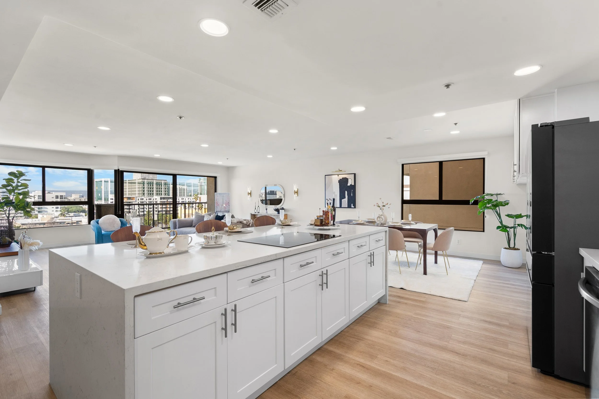 Bright and modern open-concept kitchen and living space with a white island, dining table, and large windows showing a cityscape view.