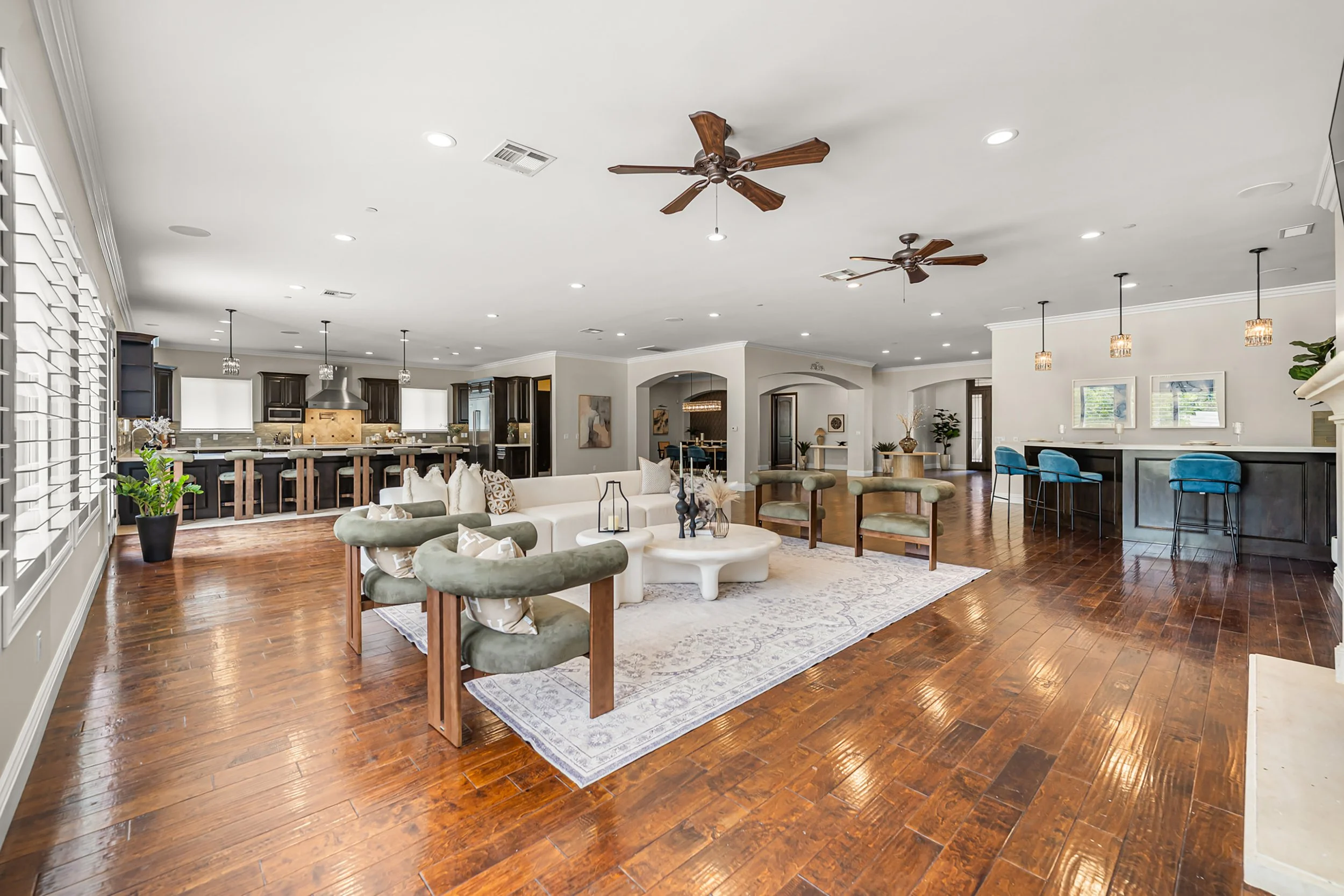 Open-concept living room and kitchen with hardwood floors, white sofa, green chairs, wooden bar stools, dark kitchen cabinets, stainless steel appliances, ceiling fans, pendant lights, large windows, and decorative items.
