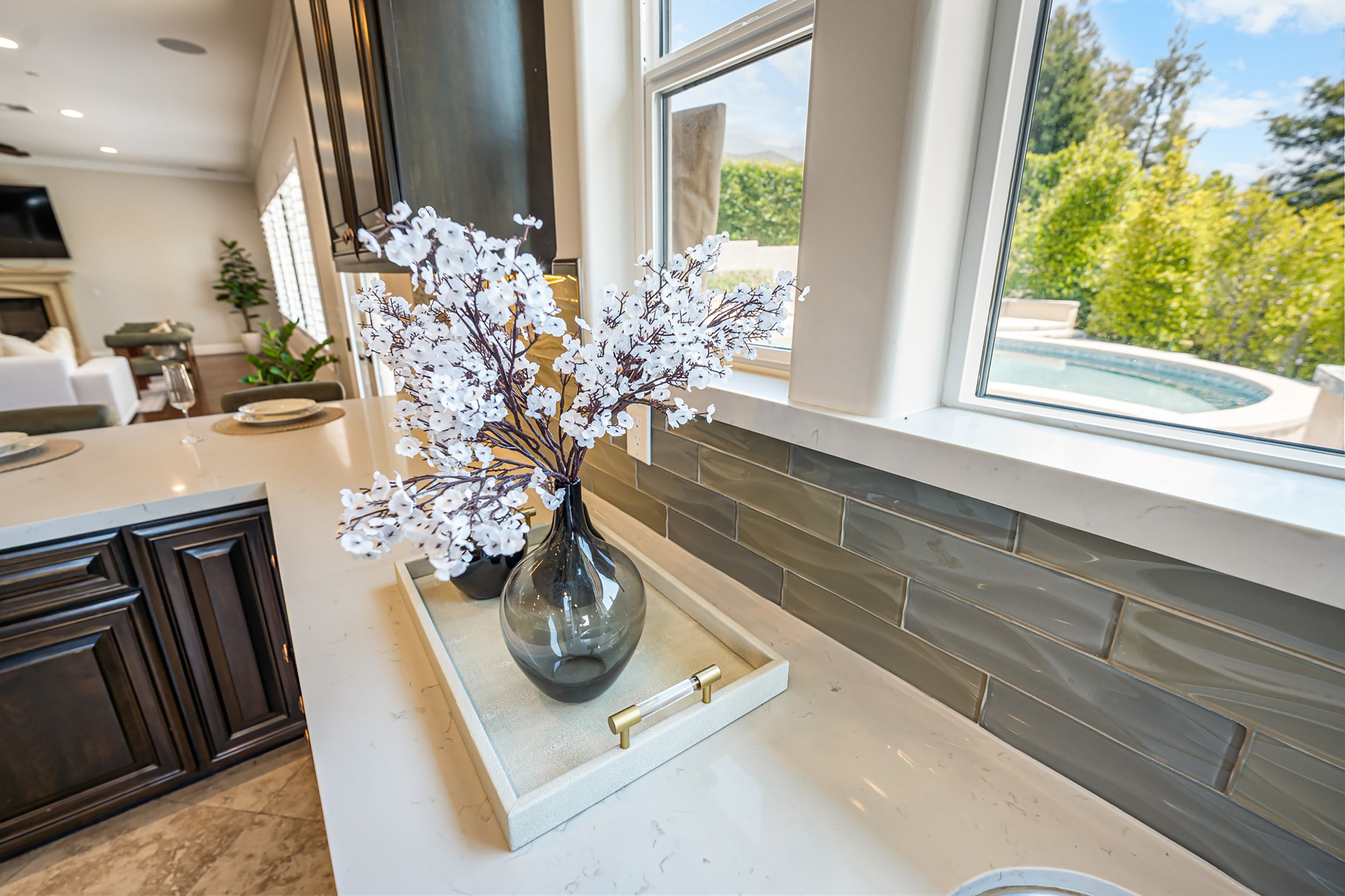 A kitchen windowsill with a flower arrangement in a glass vase, kitchen cabinets, and a view of greenery and a pool outside through the window.