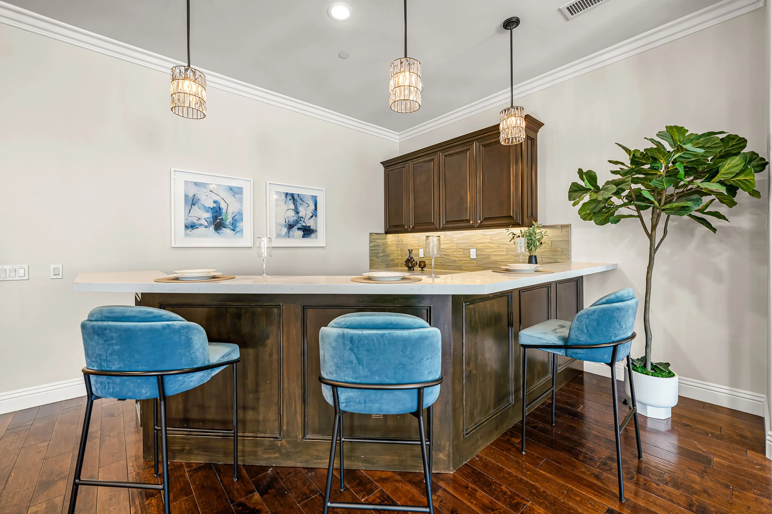 A modern kitchen and dining area with a white countertop island, blue velvet chairs, dark wooden cabinets, abstract blue artwork on the wall, pendant lights, a green plant, and dark hardwood flooring.