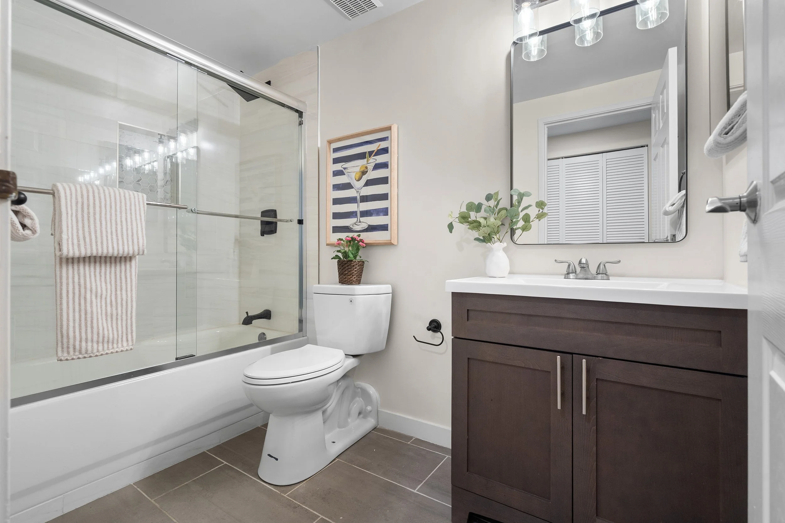 Modern bathroom with a bathtub and shower, toilet, dark wood vanity, and large mirror. Decor includes framed artwork of a martini glass, potted plant, and folded towels.
