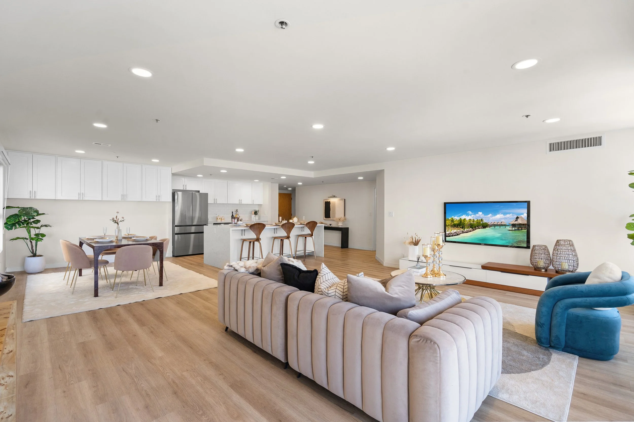 Spacious open-plan living room with white cabinets, wooden floors, cream sofas, a blue armchair, a dining table with chairs, and a large flat-screen TV displaying a beach scene.