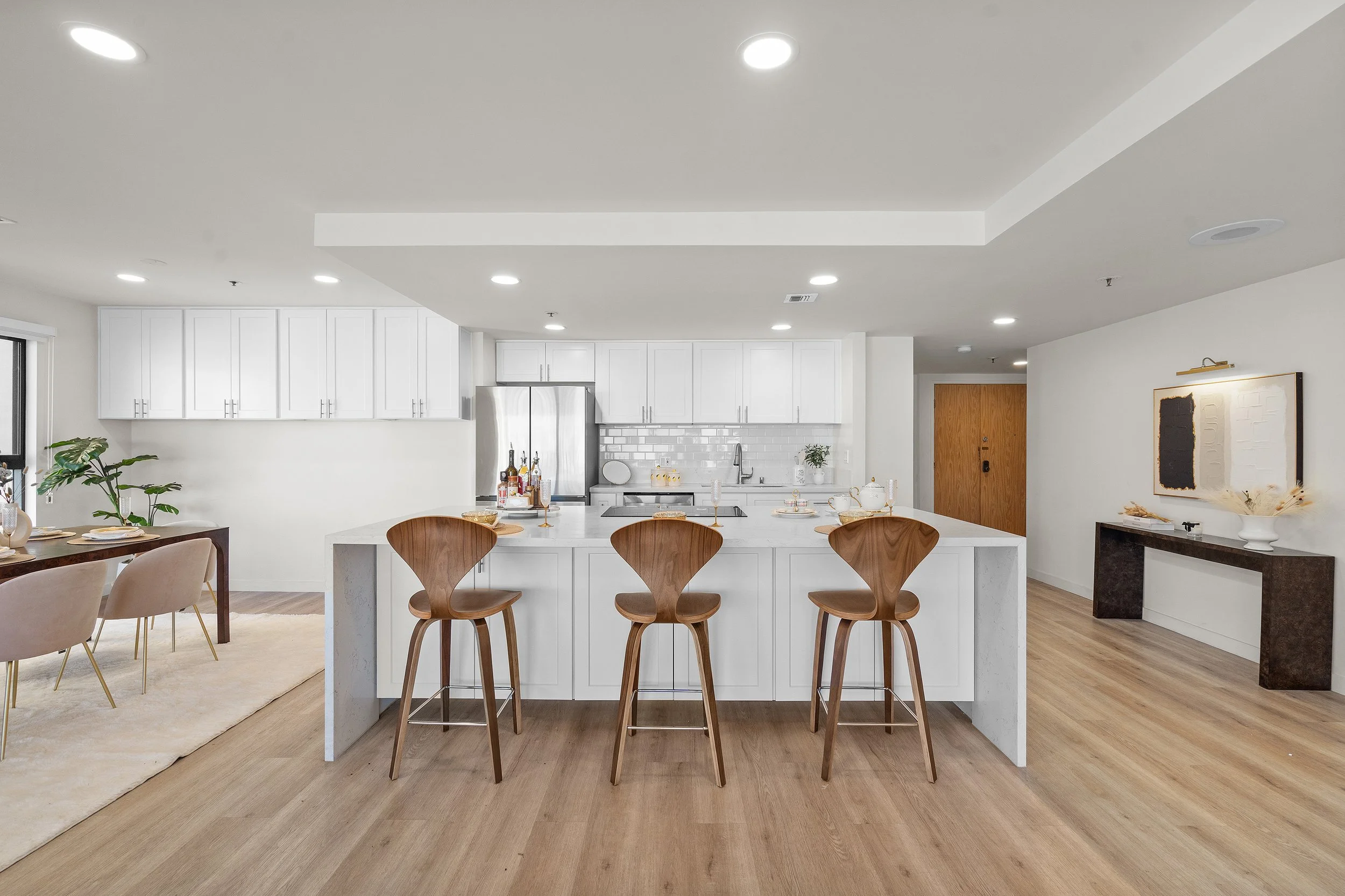 Modern kitchen and dining area with white cabinets, a central island with wooden bar stools, and a dining table with beige chairs, hardwood flooring, and recessed lighting.