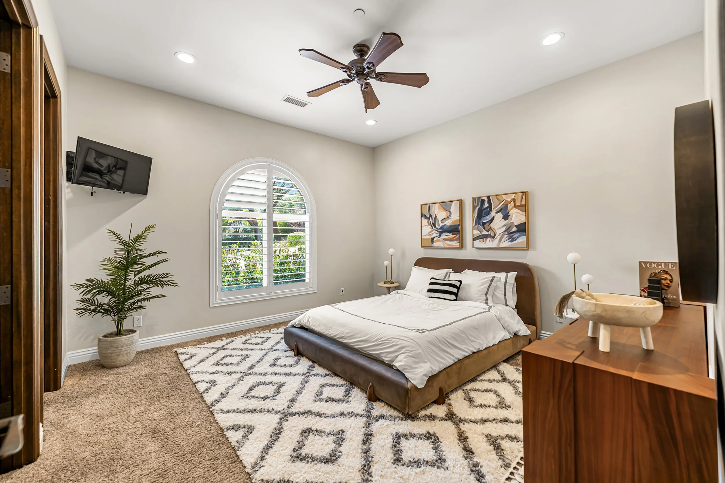 A cozy bedroom with a large bed featuring white bedding and a striped pillow, a potted plant near the window, wall art above the bed, a wall-mounted flat-screen TV, a ceiling fan, and decorative lamps, with natural light coming through arched window 