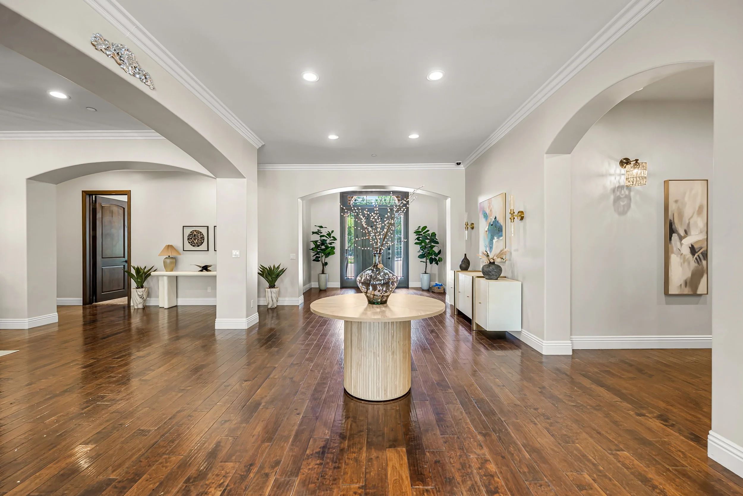 Spacious, modern living room with hardwood floors, white walls, arched entryways, and contemporary art. Decor includes plants, paintings, and a round wooden table with a decorative glass vase.
