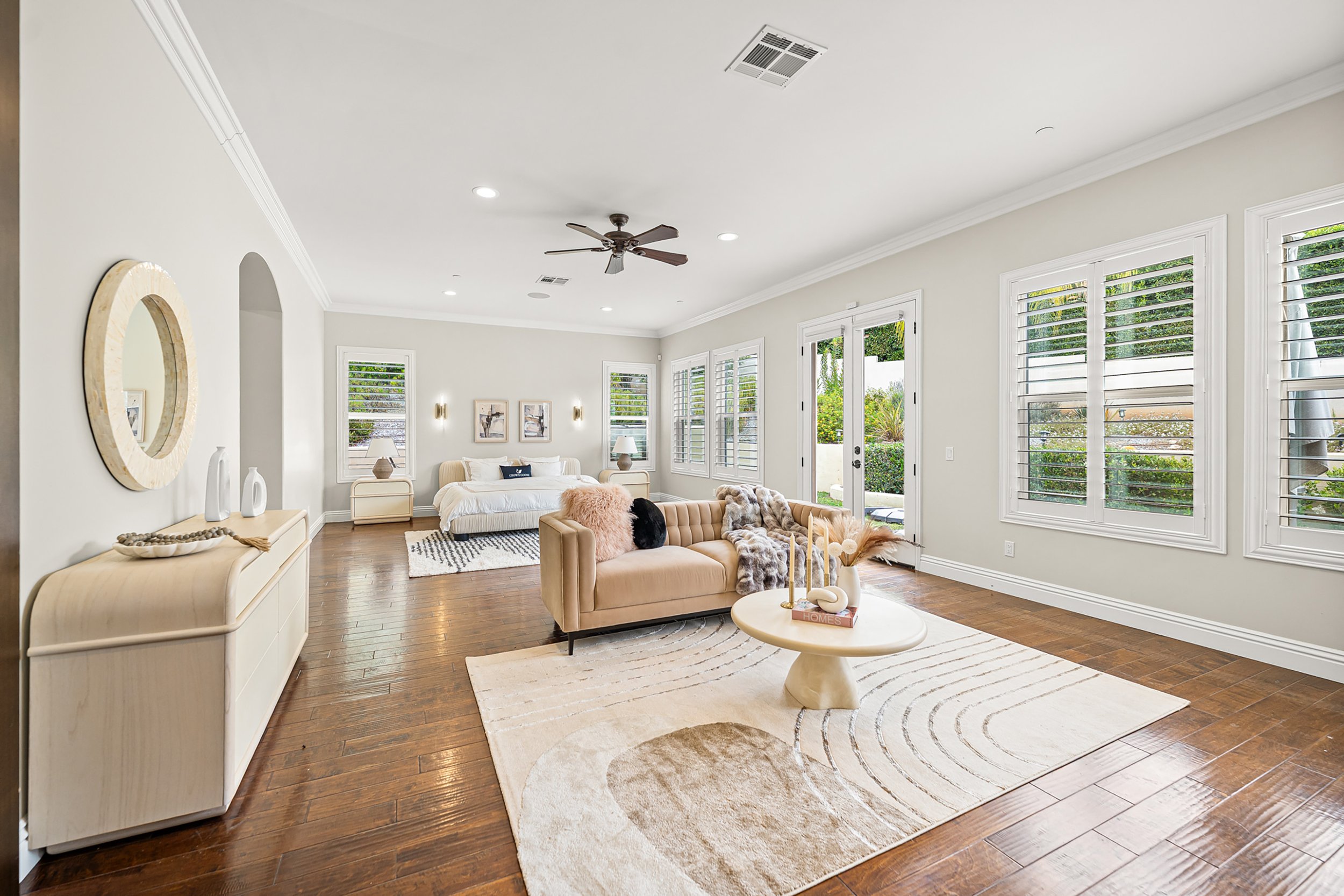 Bright living room with large windows, white walls, hardwood floors, a beige sofa, a cream-colored sideboard with decorative vases, wall art, a white armchair, and a round coffee table with decorative items.