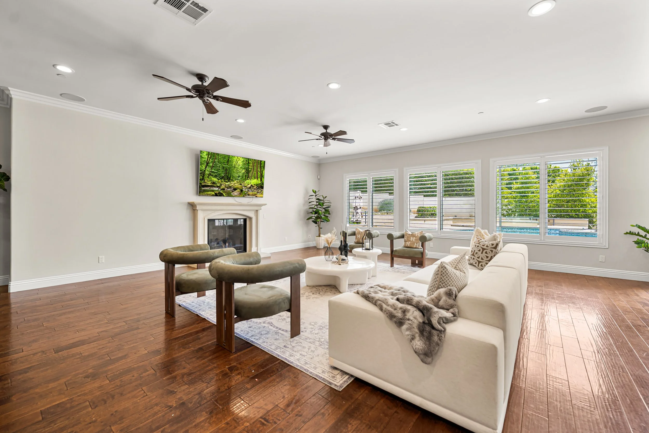 Bright living room with white walls, hardwood floor, a fireplace, a wall-mounted TV showing a forest scene, large windows with white blinds, a beige sofa decorated with patterned cushions and a faux fur throw, a round white coffee table, and potted p