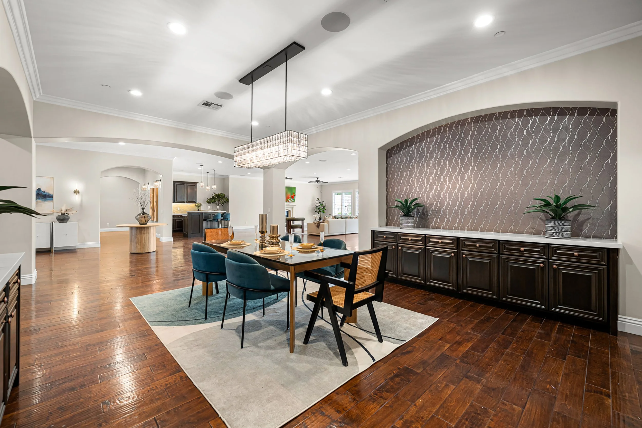 A spacious, modern dining and living area with hardwood floors, white walls, and a mix of contemporary and traditional furniture. There is a dining table with blue chairs, various decorative items, potted plants, and a kitchen area in the background 