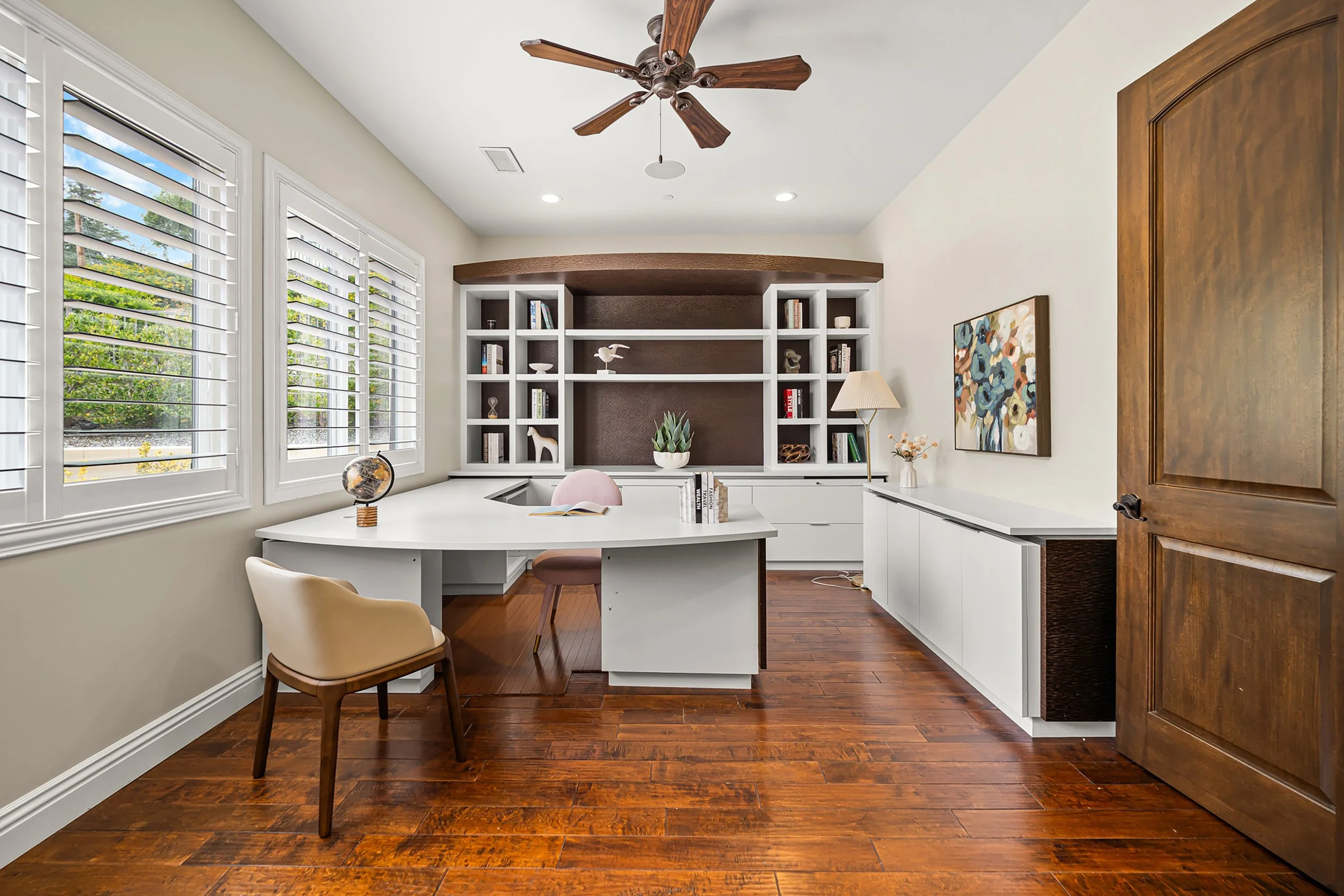 A home office with a white desk, beige and pink chairs, a globe, open book, and decorative objects. White built-in shelves with plants, books, and art are on the back wall. Large windows with white plantation shutters let in natural light. Hardwood f