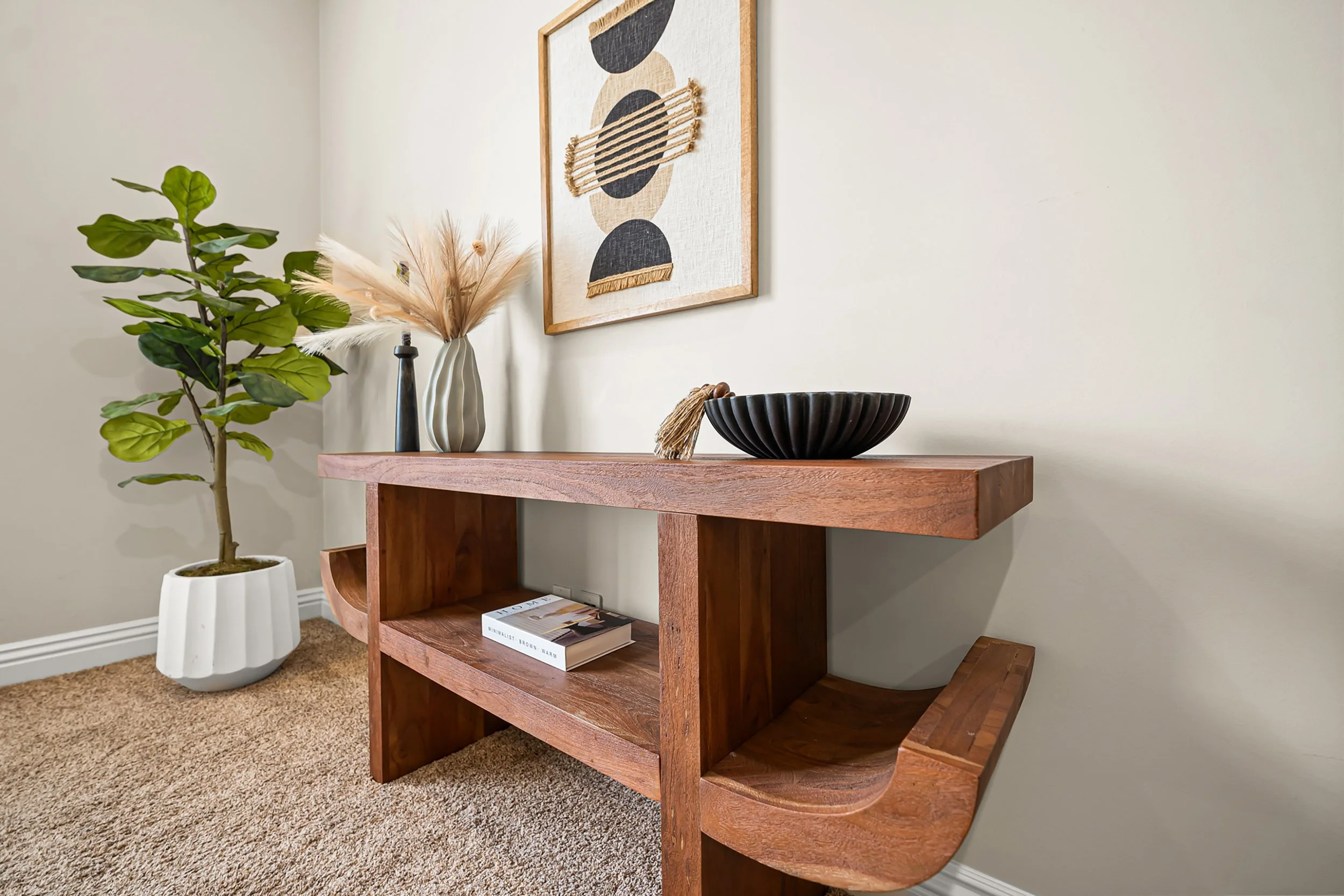 Decorative console table in a living room with a large potted plant, a vase with dried pampas grass, a black bowl, and a magazine, with an abstract framed wall art piece hanging above.