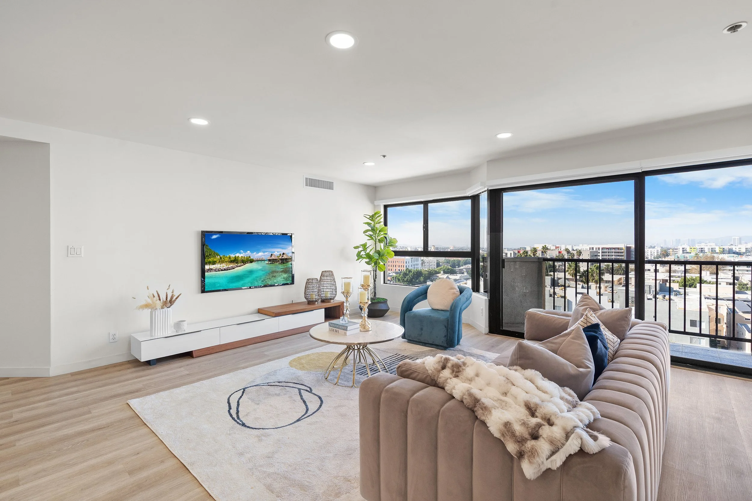 Living room with beige sofa, blue armchair, white TV stand, wall-mounted TV showing beach scene, large windows with city view, light wood floors, rug, decorative items, indoor plant, and beige curtains.