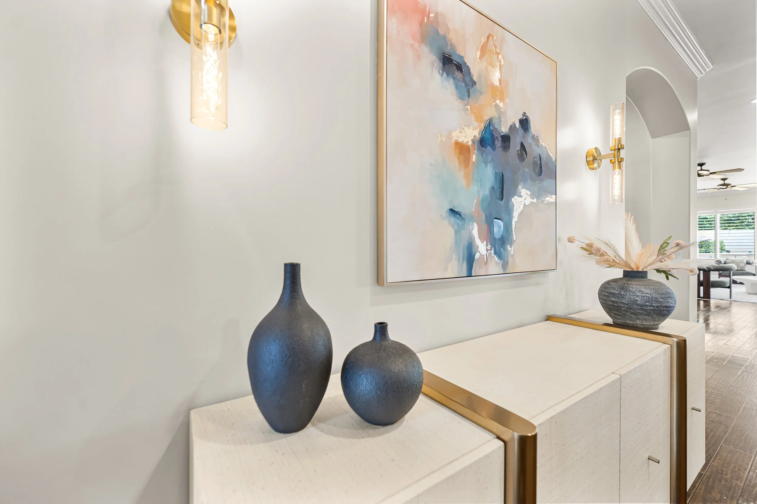 A modern living room with abstract wall art, decorative vases on a white sideboard, and a vase with dried flowers. Gold accents are present in the lighting fixtures and sideboard trim.