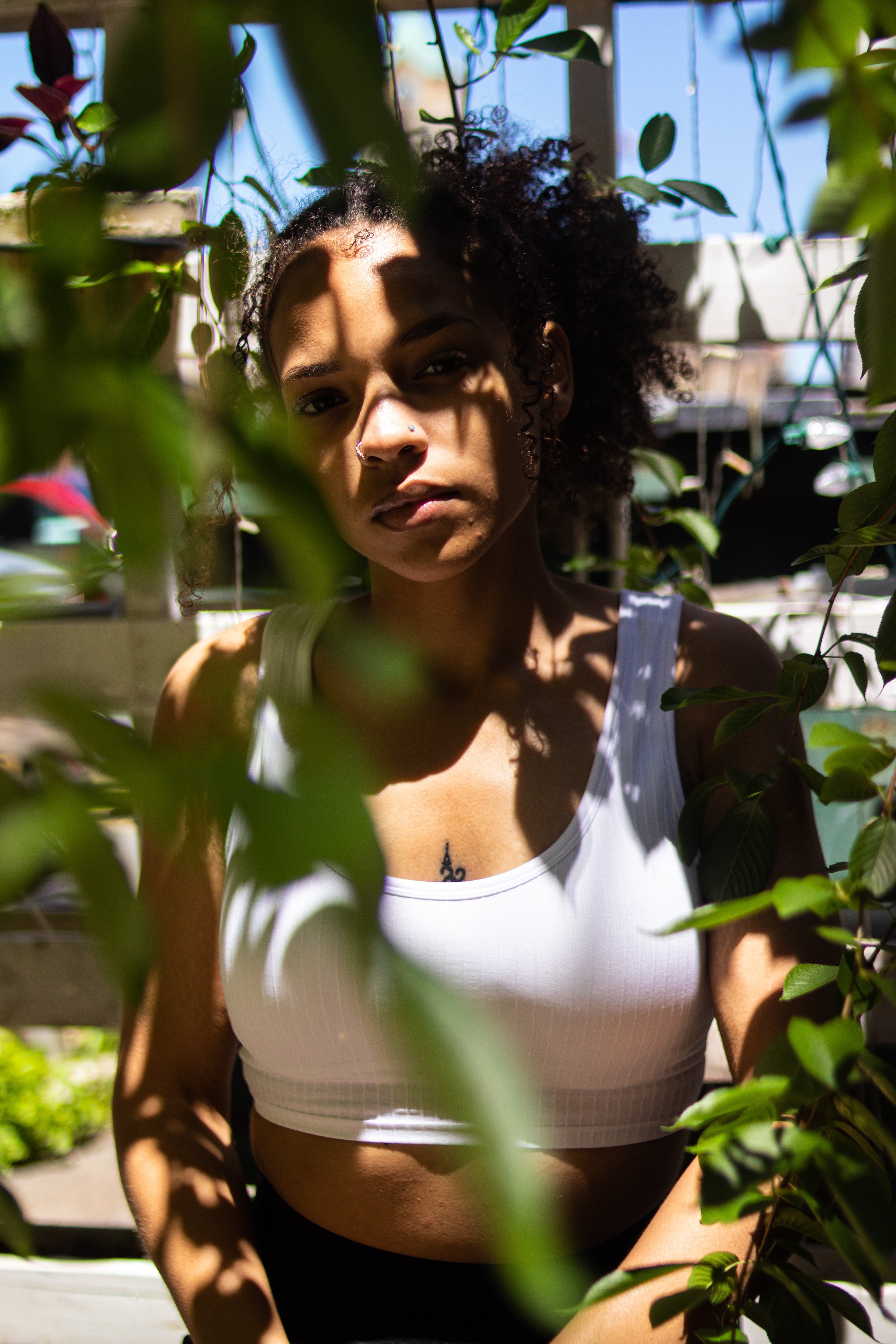 A young woman with curly hair and a nose piercing gazes through green foliage with a serious expression, wearing a white tank top with sunlight casting shadows on her face.