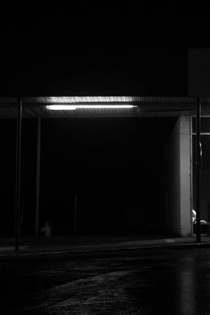 A dark, night-time scene showing a bus stop shelter with an illuminated light on the ceiling. The bus stop is on a street with a wet road surface, and there are supporting poles and a building corner visible.