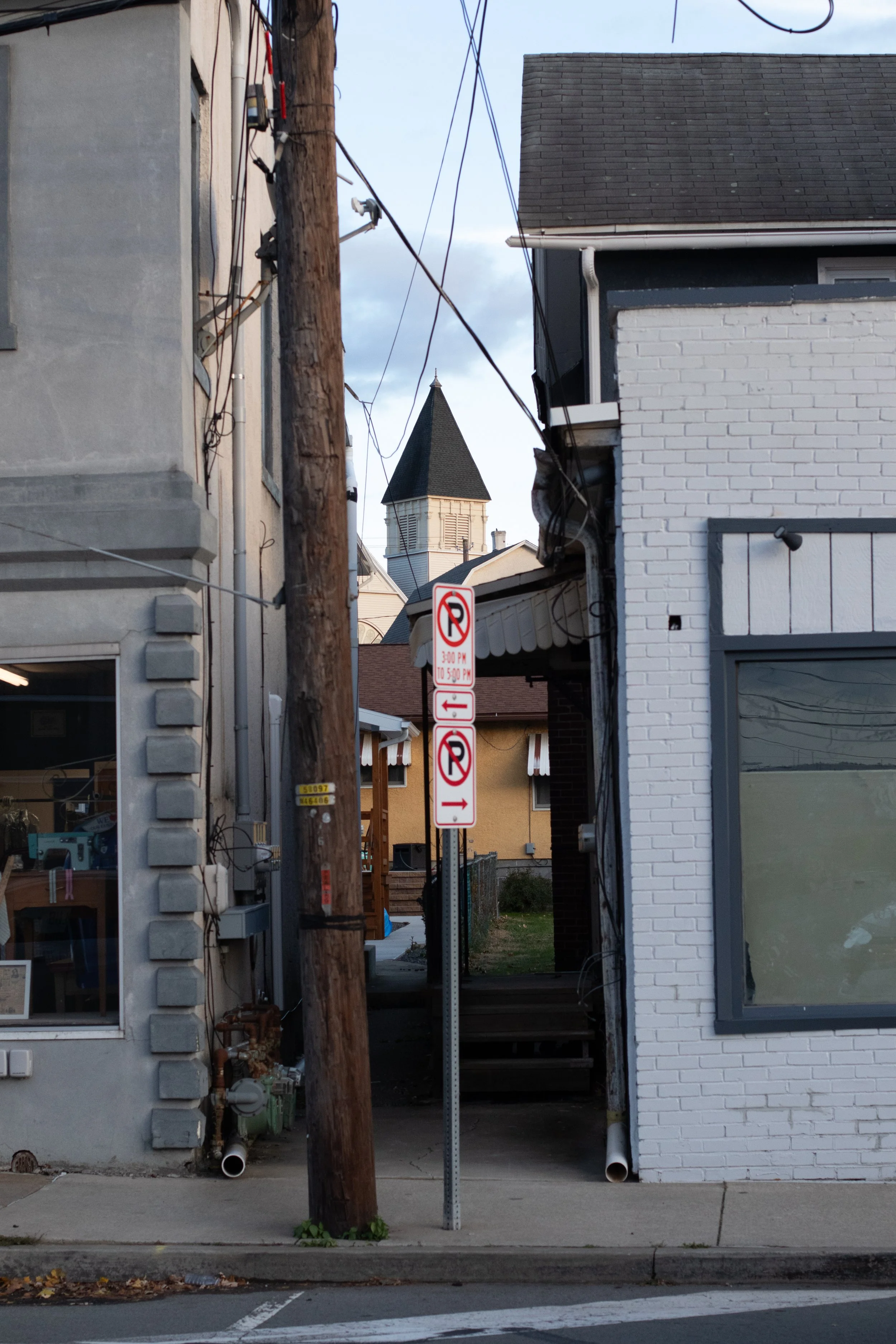 Street scene with a utility pole and two signs indicating no parking in both directions, a church steeple visible in the background, and the facades of two buildings, one with white brick and the other with gray concrete.