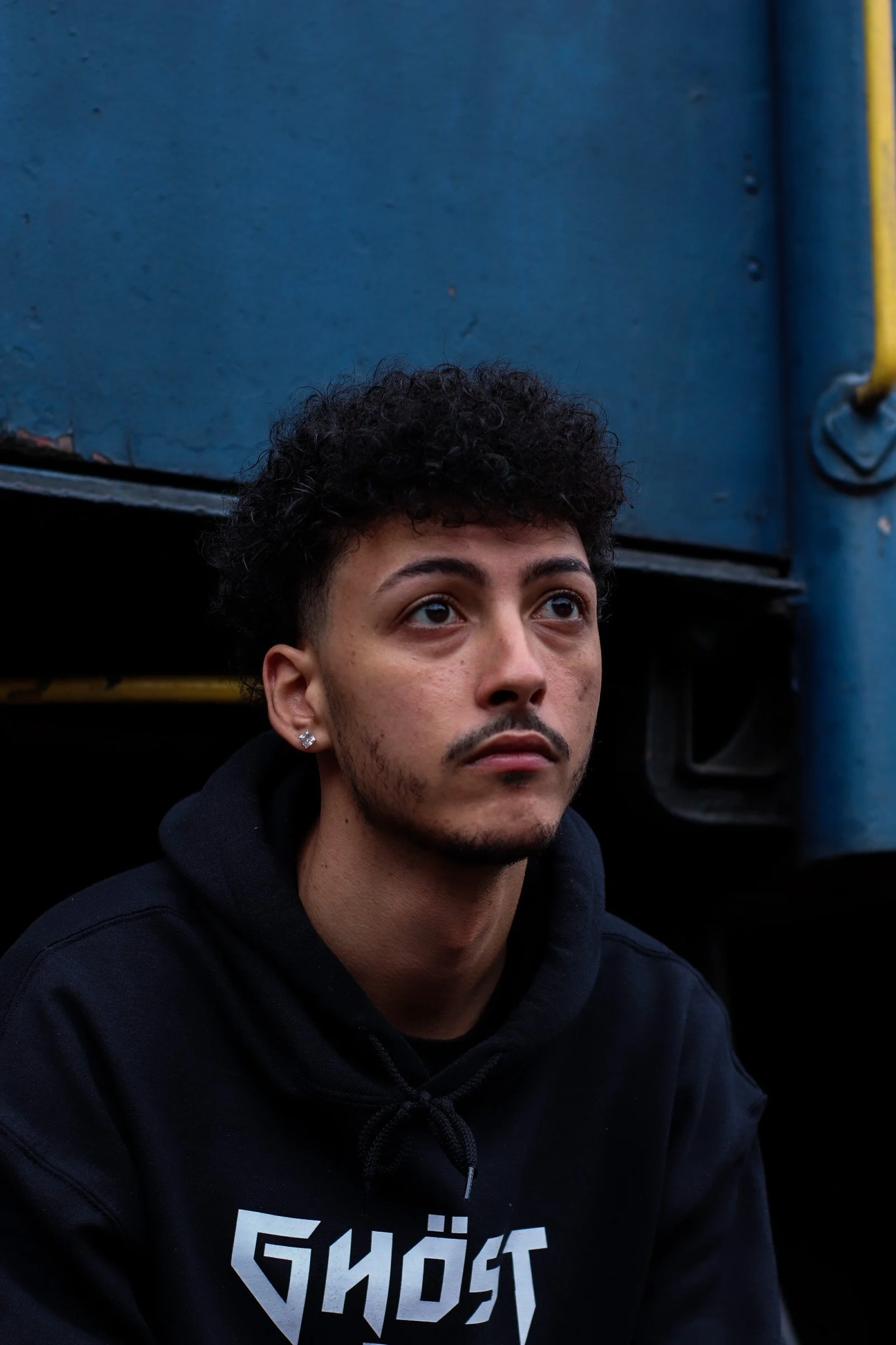 A young man with curly black hair and earrings, wearing a black hoodie with the word "GHOST" on it, looking upward outdoors near a blue structure.