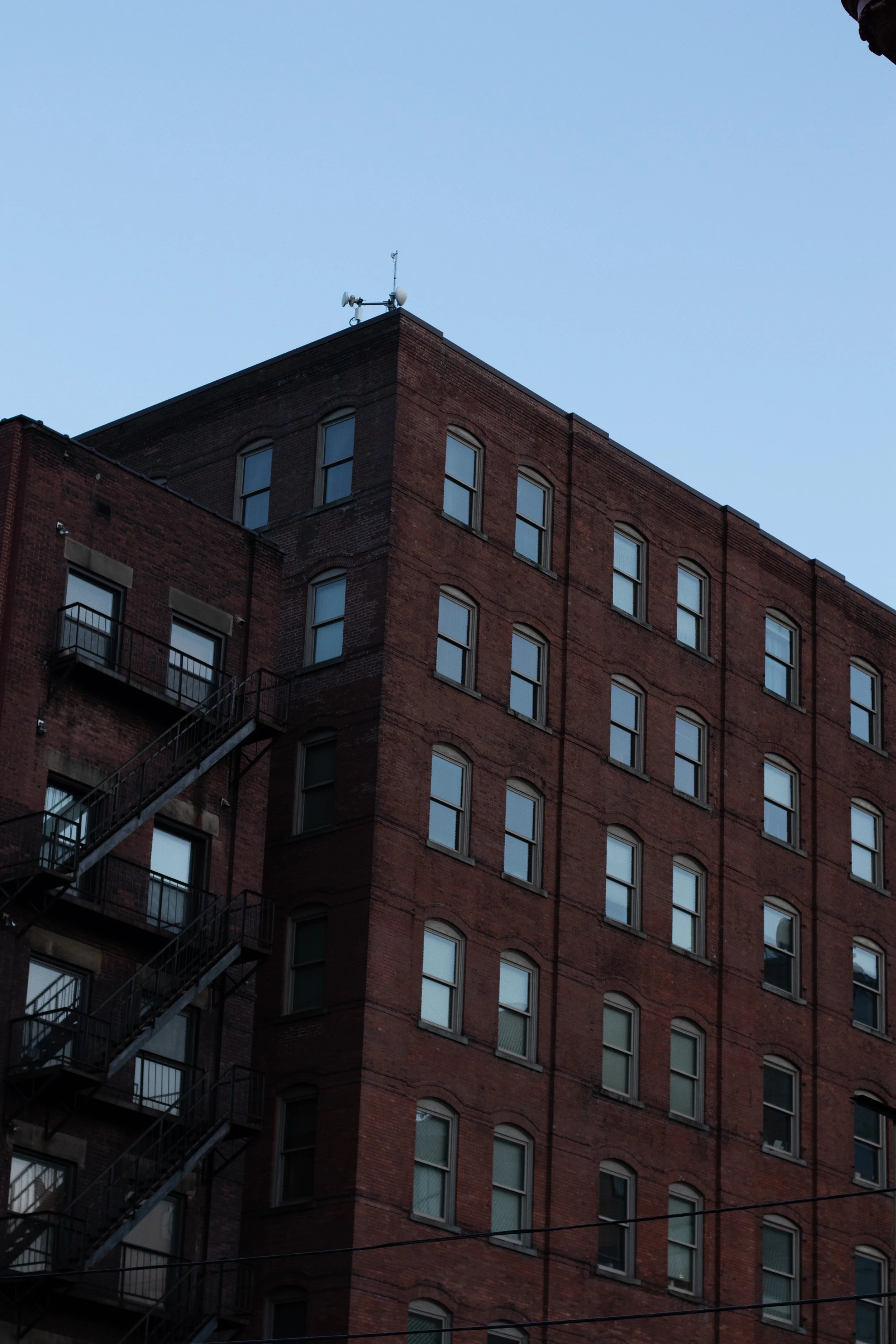 A tall, red brick building with multiple windows and an external fire escape against a clear blue sky.