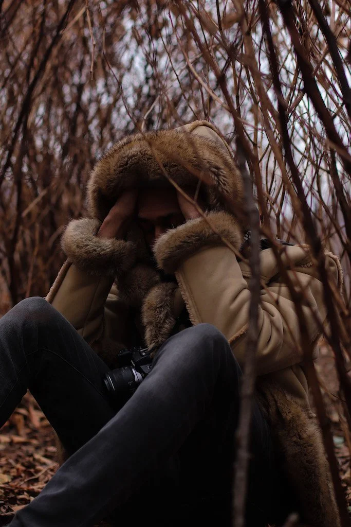 Person sitting among dry, brown foliage, wearing a tan coat with a fur-lined hood, partially covering their face with hands.
