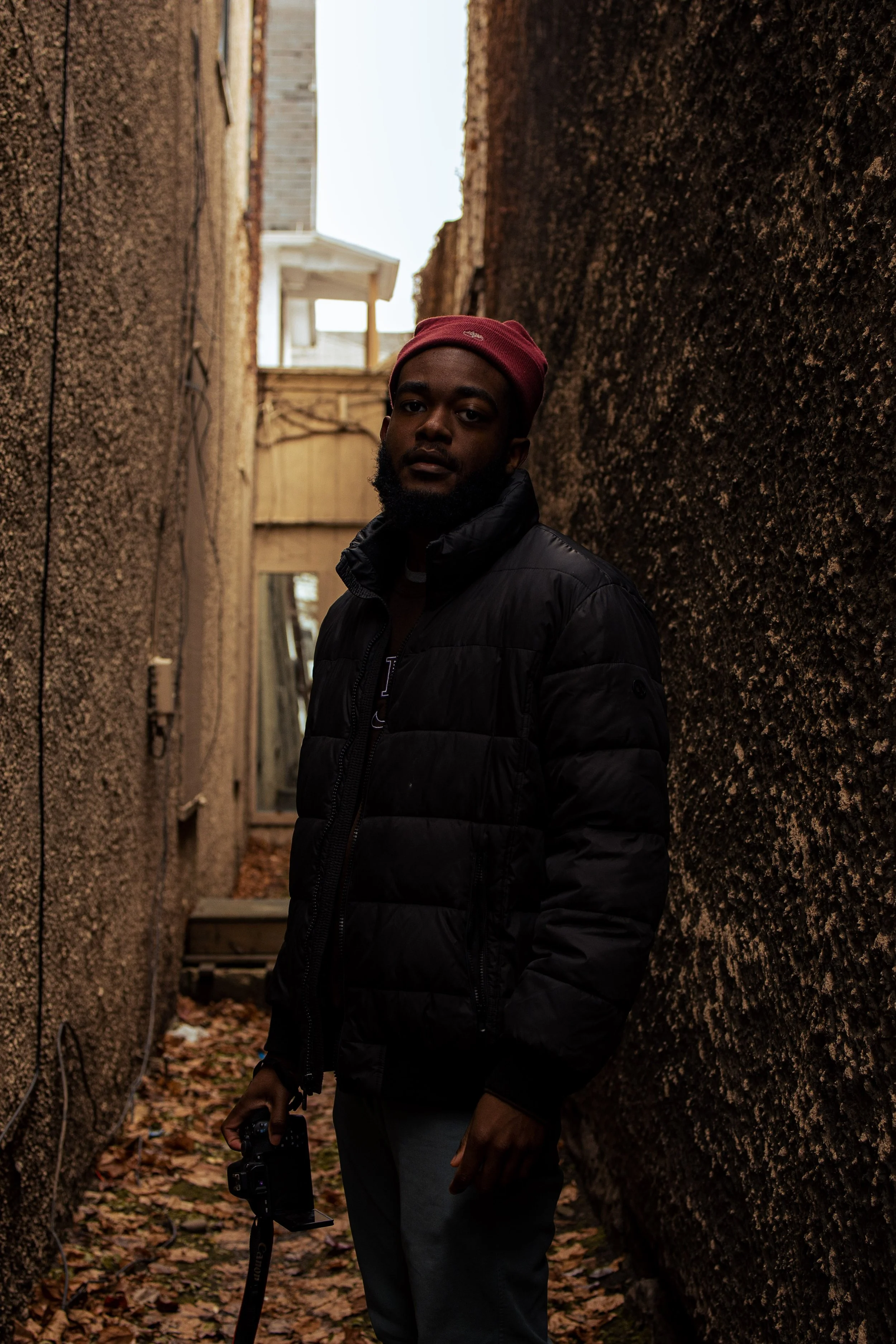 A man with a red beanie and black jacket standing in a narrow alley with brown textured walls, holding a camera in his right hand.