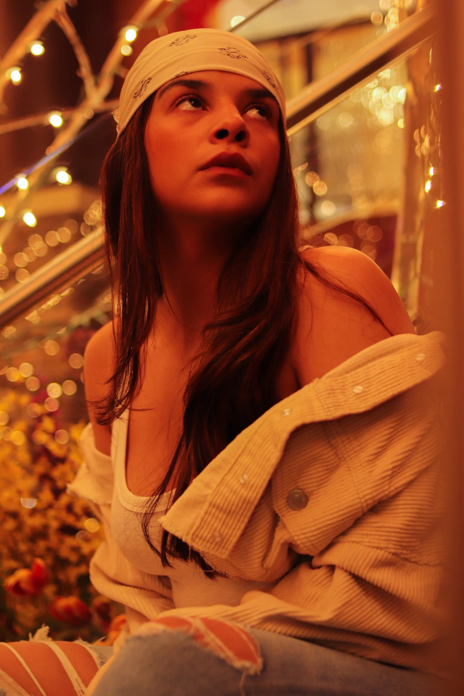 A young woman with long brown hair, wearing a beige bandana and off-shoulder jacket, sits in front of a glass surface with warm string lights and blurred floral background.