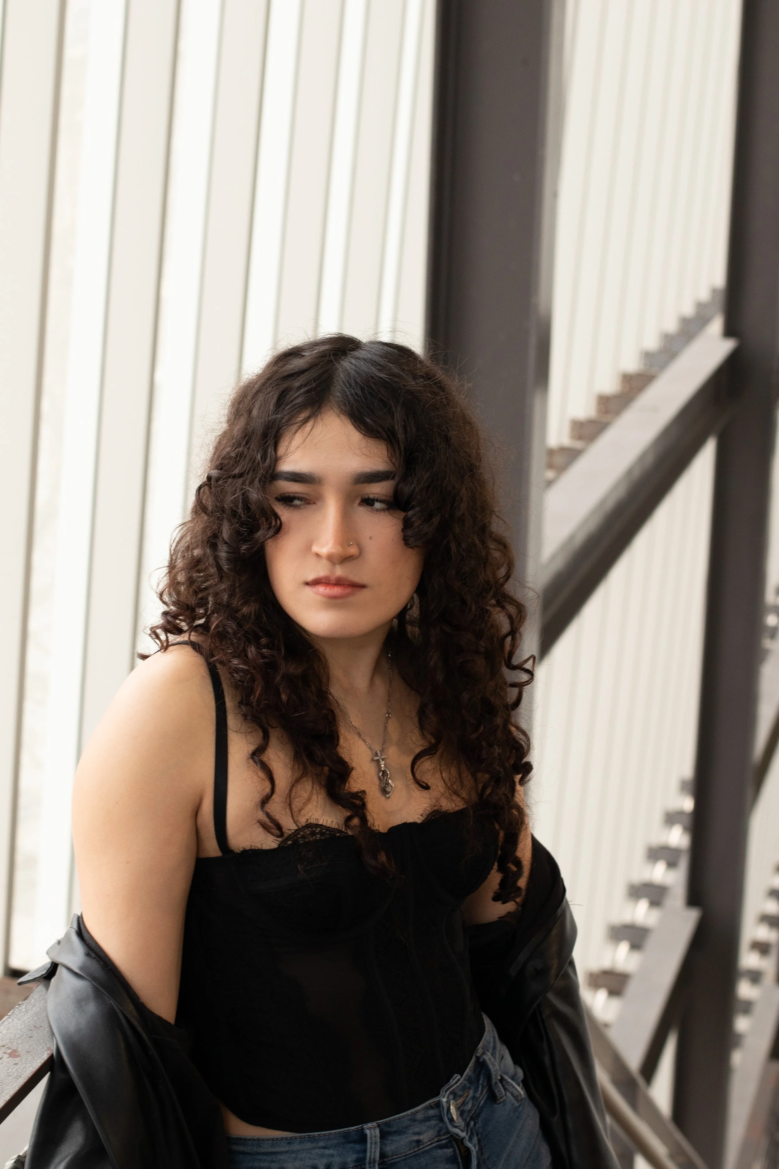 A young woman with curly brown hair wearing a black tank top, a necklace, and a leather jacket draped over her arms, standing indoors near a staircase with white vertical paneling in the background.