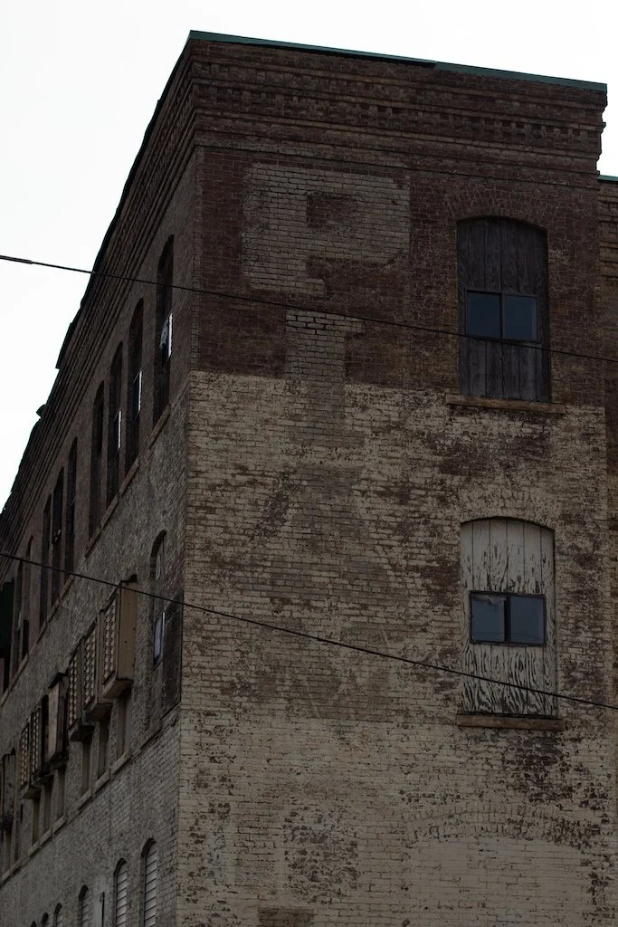 A large, old brick building with two visible windows and vandalized boarded-up windows. There are two cables crossing the image, and a faint outline of a person holding a balloon is seen at the top.