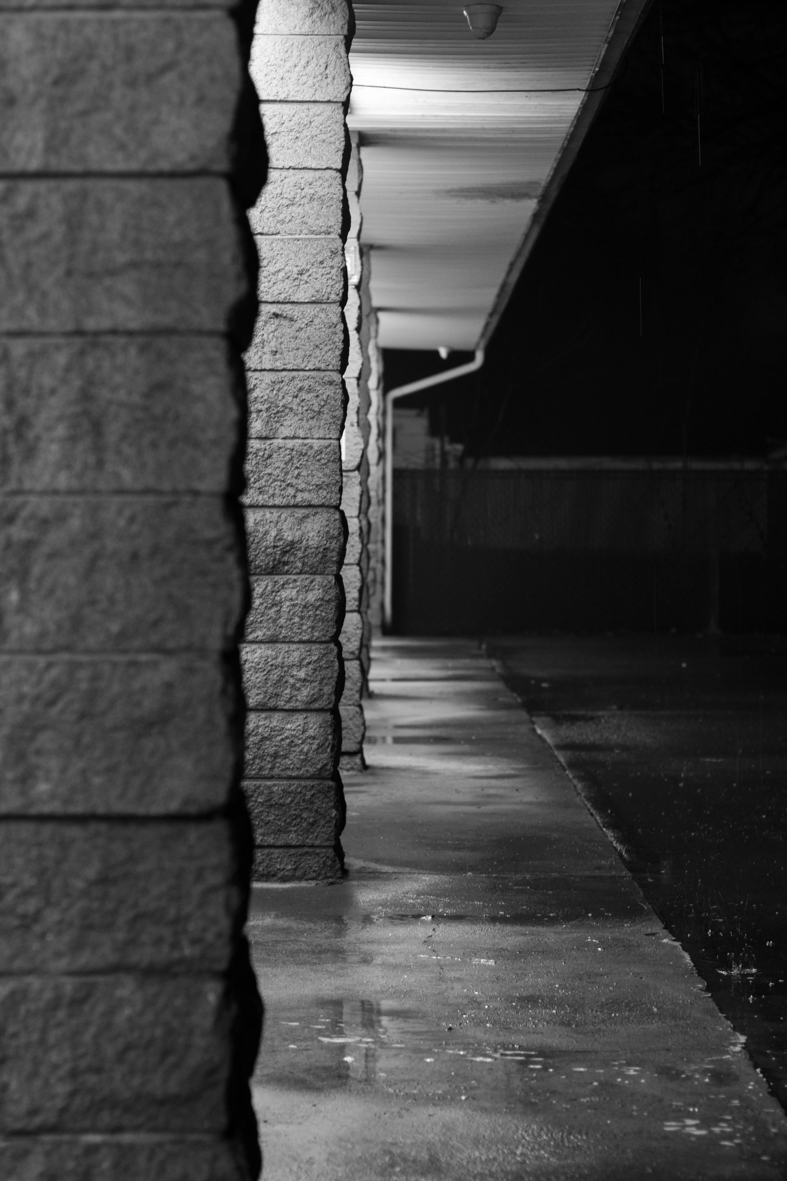 Black and white photo of a row of brick columns and a covered walkway at night, with reflections on wet pavement.