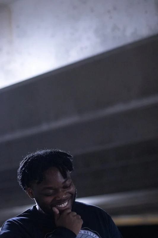 A young man with dreadlocks smiling and holding his hand to his chin, standing under a bridge or overpass.