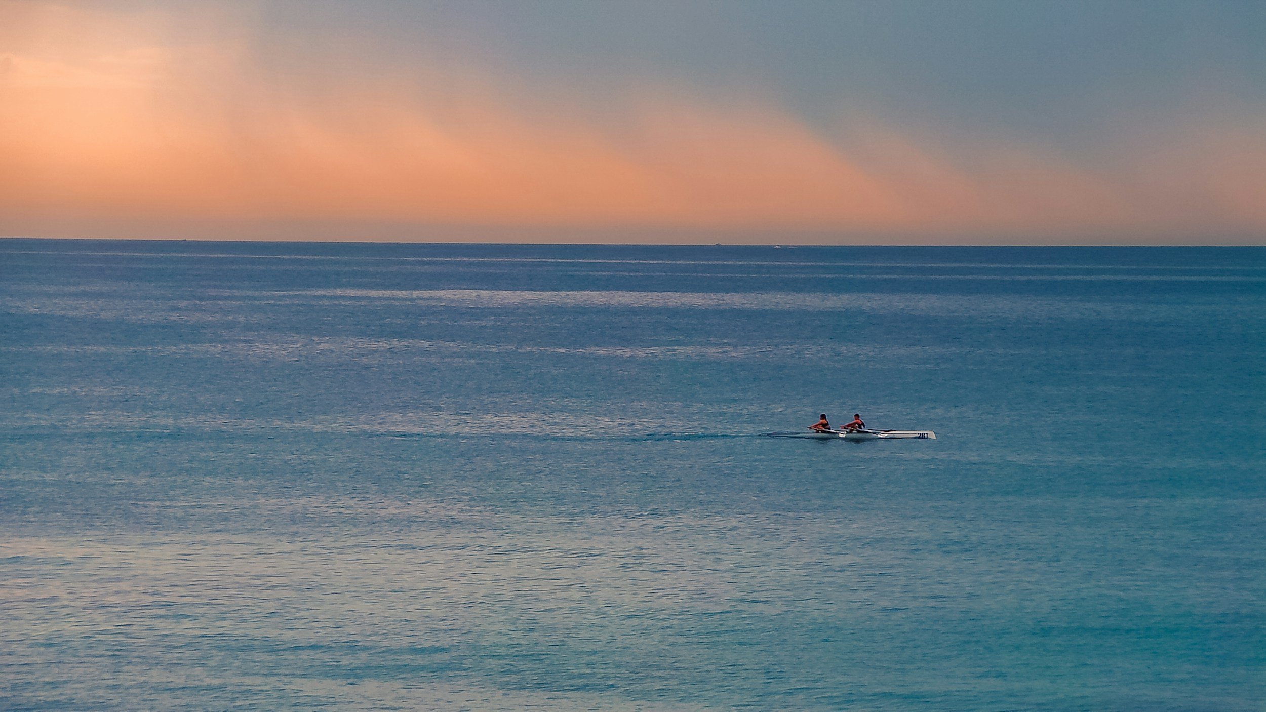 Two people in a tandem ocean rowing boat rowing on calm ocean water during sunset with a colorful sky.