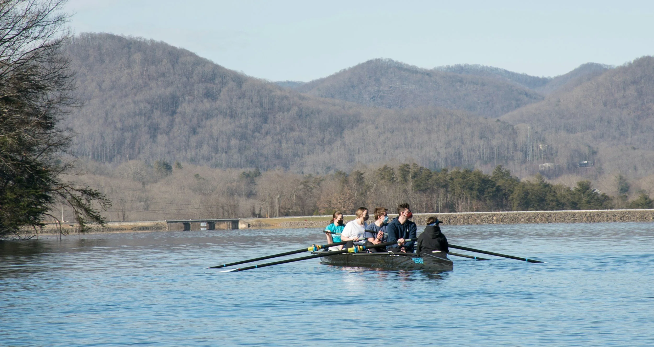 A group of five people in a rowboat on a calm lake with mountains in the background.