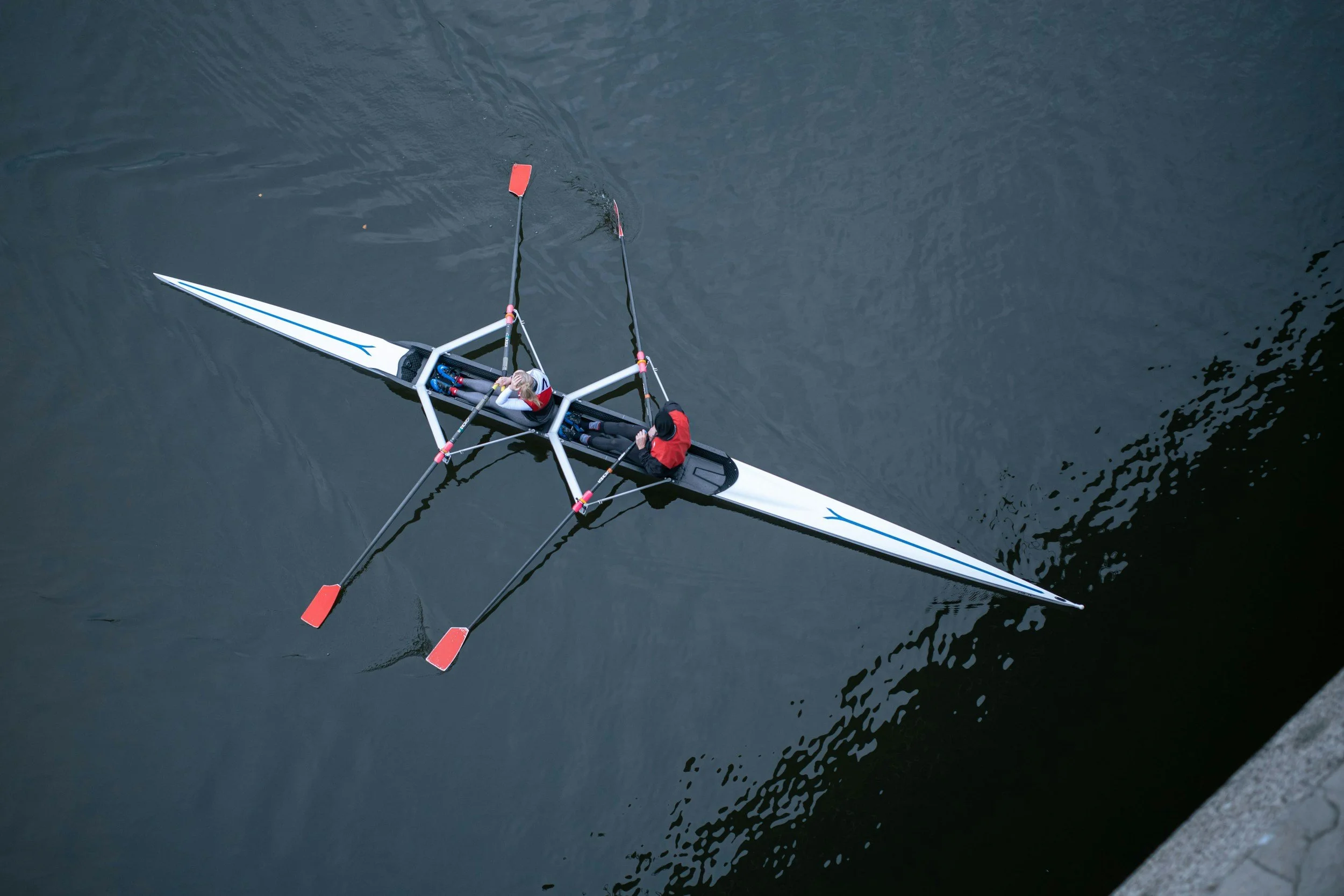Aerial view of two people in a rowing shell on dark water near a concrete edge.
