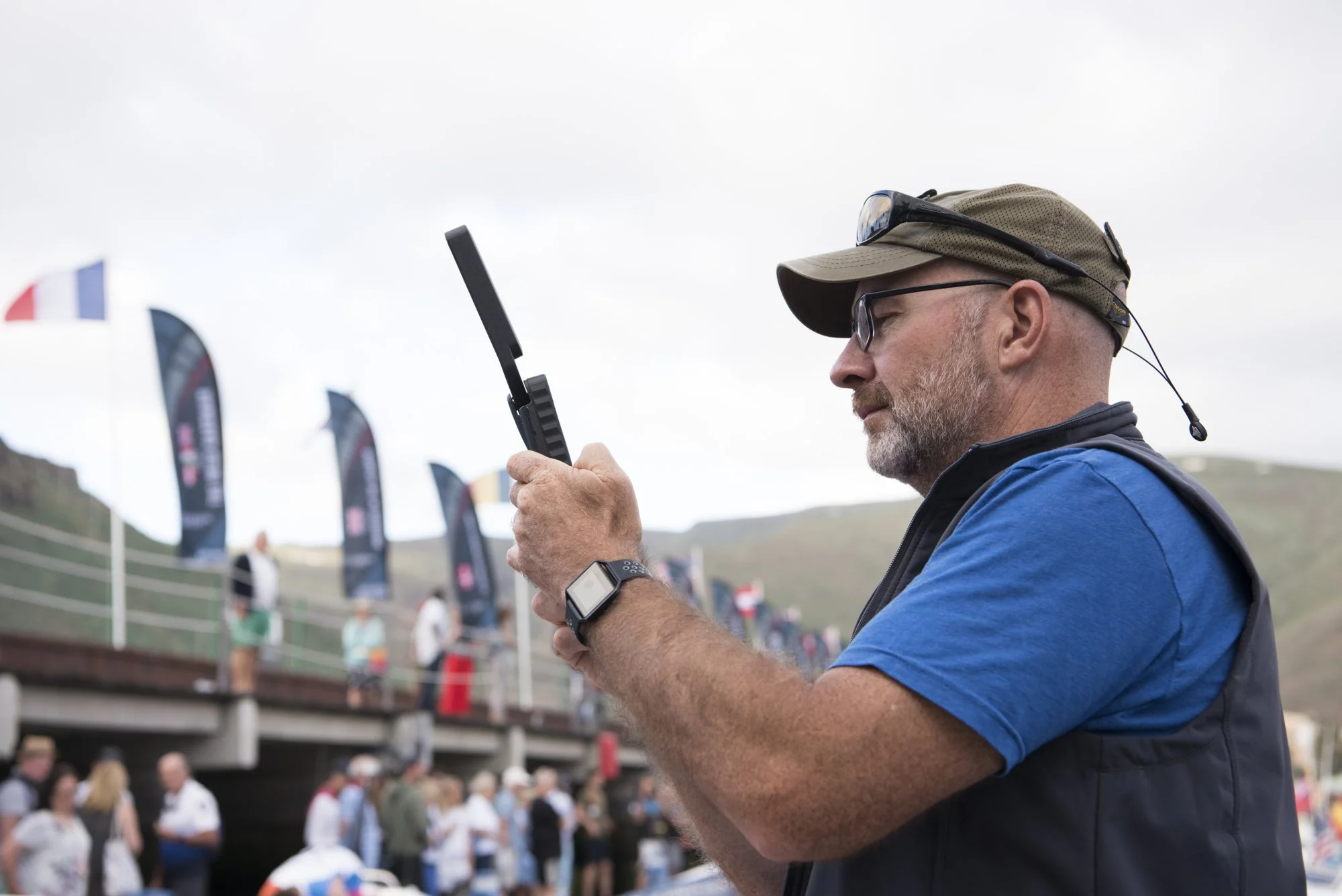 A man wearing glasses, a cap, and a blue shirt, holding a satellite-phone, standing outdoors at an event with flags and people in the background.